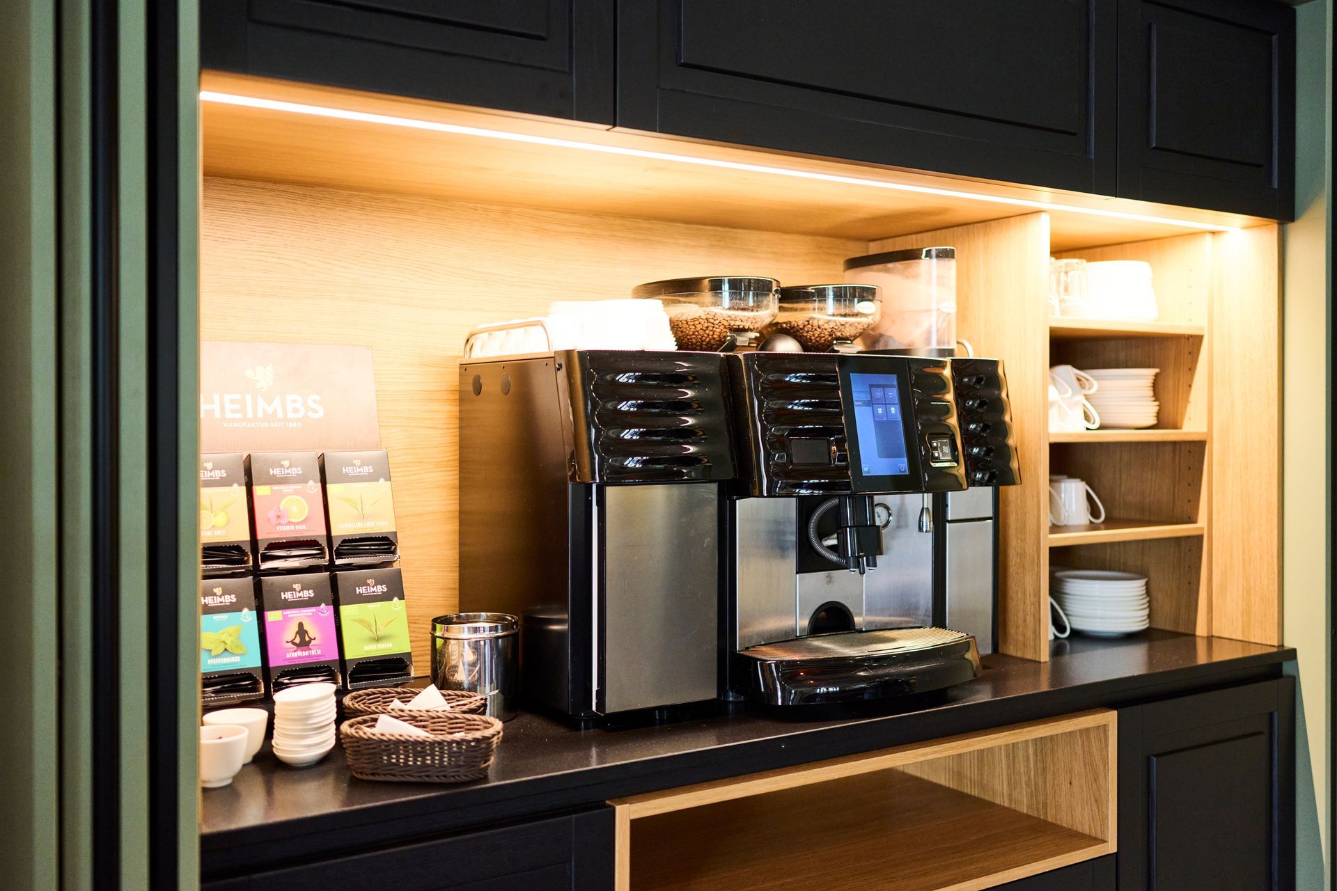 Eine Kaffeestation mit einer professionellen Espressomaschine, verschiedenen Teebeuteln und gestapelten Tassen in einem beleuchteten Holzschrank.
