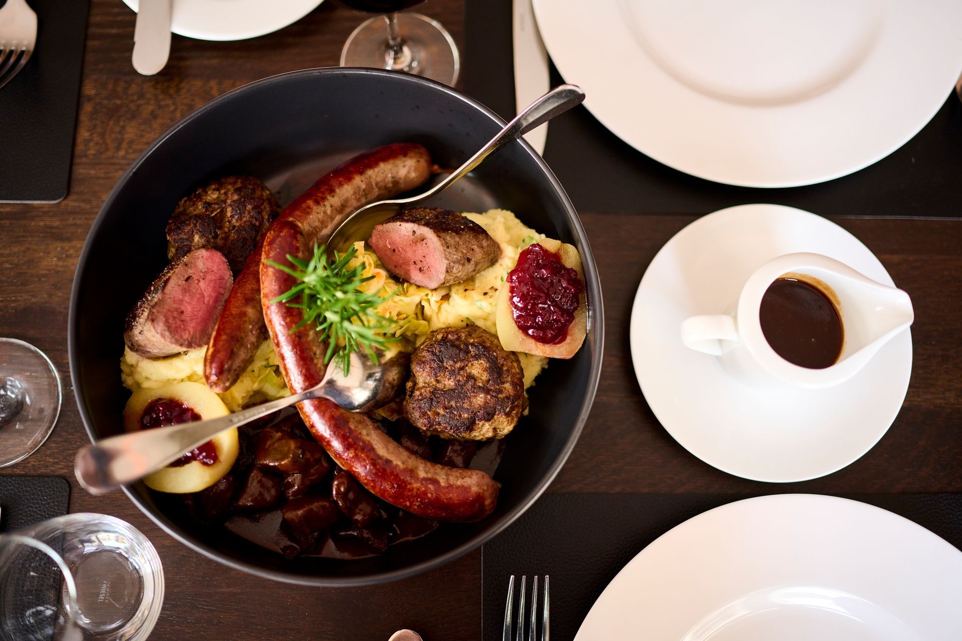 Eine große Servierschüssel gefüllt mit Wurst, Fleischbällchen, Kartoffelpüree und roter Beerenmarmelade, dazu ein Schälchen Soße.