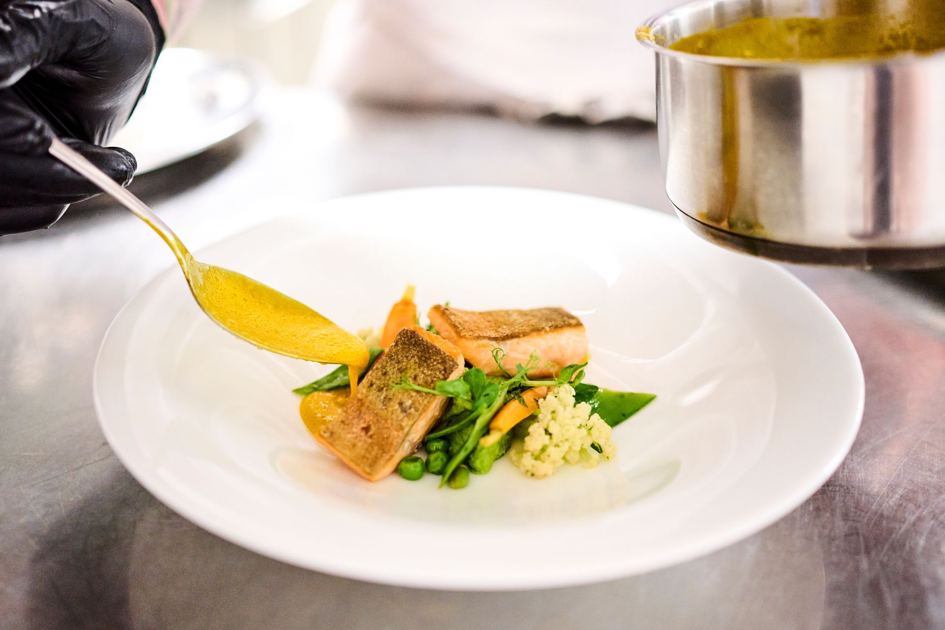 Ein Koch mit schwarzem Handschuh träufelt mit einem goldenen Löffel Sauce über ein angerichtetes Gericht mit gebratenem Fisch und Gemüse.