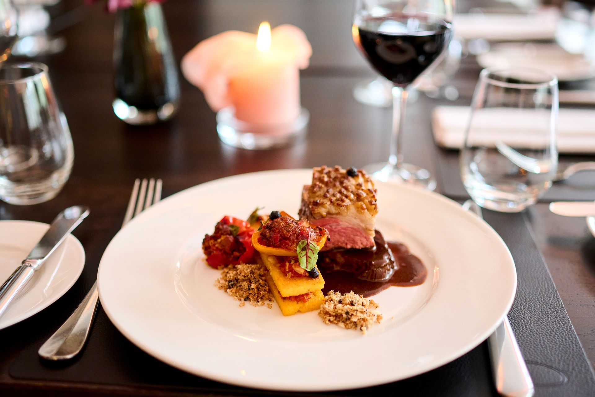 Ein Gourmet-Menü aus gebratenem Fleisch und Beilagen auf einem weißen Teller, serviert auf einem dunklen Tisch mit Wein und einer Kerze.