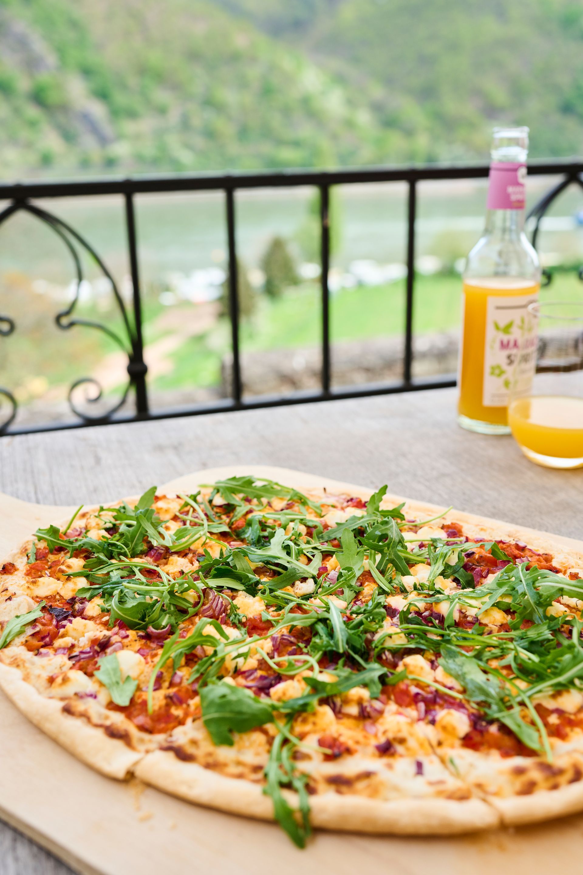 Eine Pizza mit dünnem Boden, belegt mit Rucola und Speck, auf einem Holzbrett, dazu ein Getränk in der Nähe und ein malerischer Ausblick im Hintergrund.