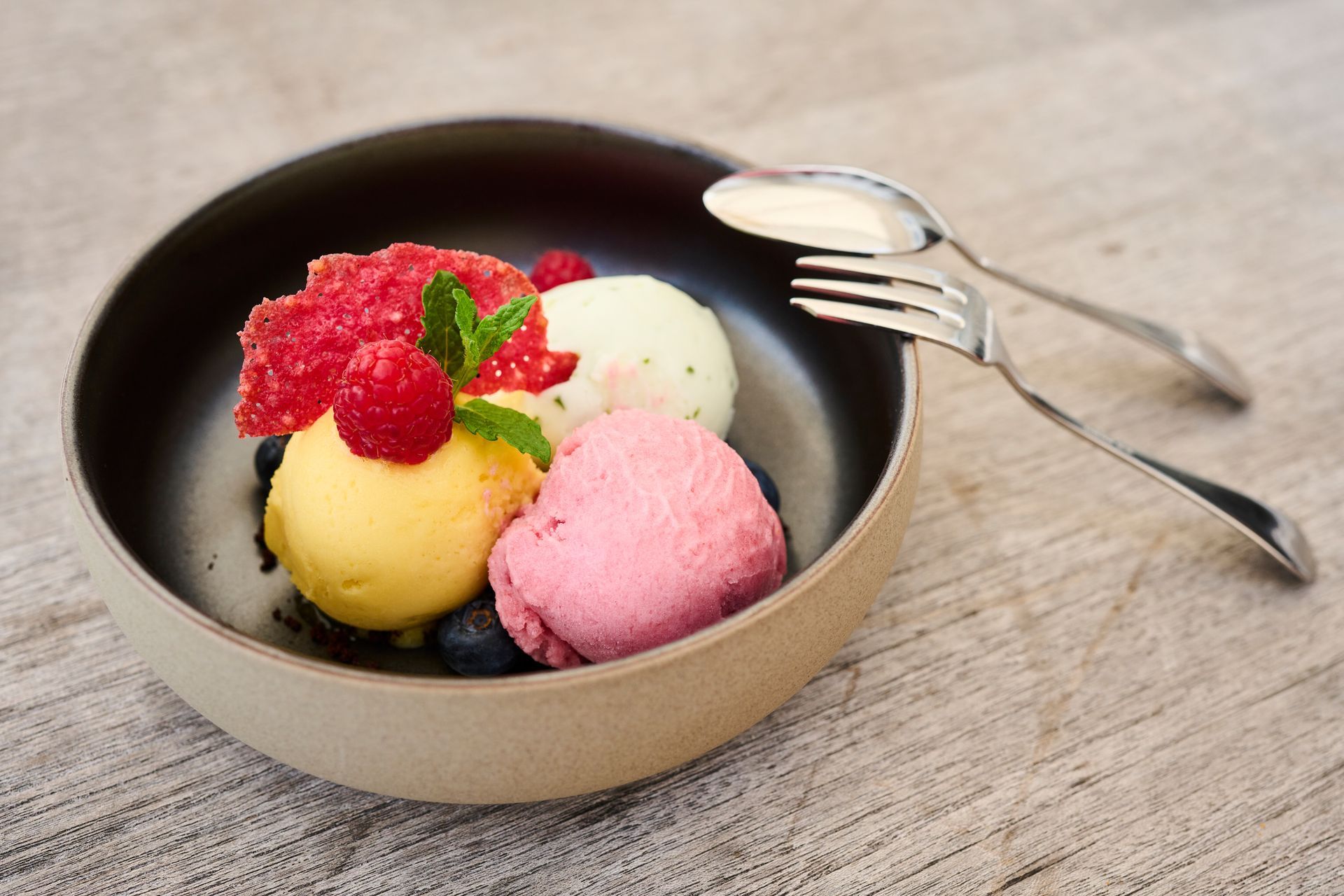 Drei Kugeln Sorbet – gelbes, rosa und weißes – in einer dunklen Schüssel mit Beeren, Minze und einem Löffel auf einer Holzoberfläche.