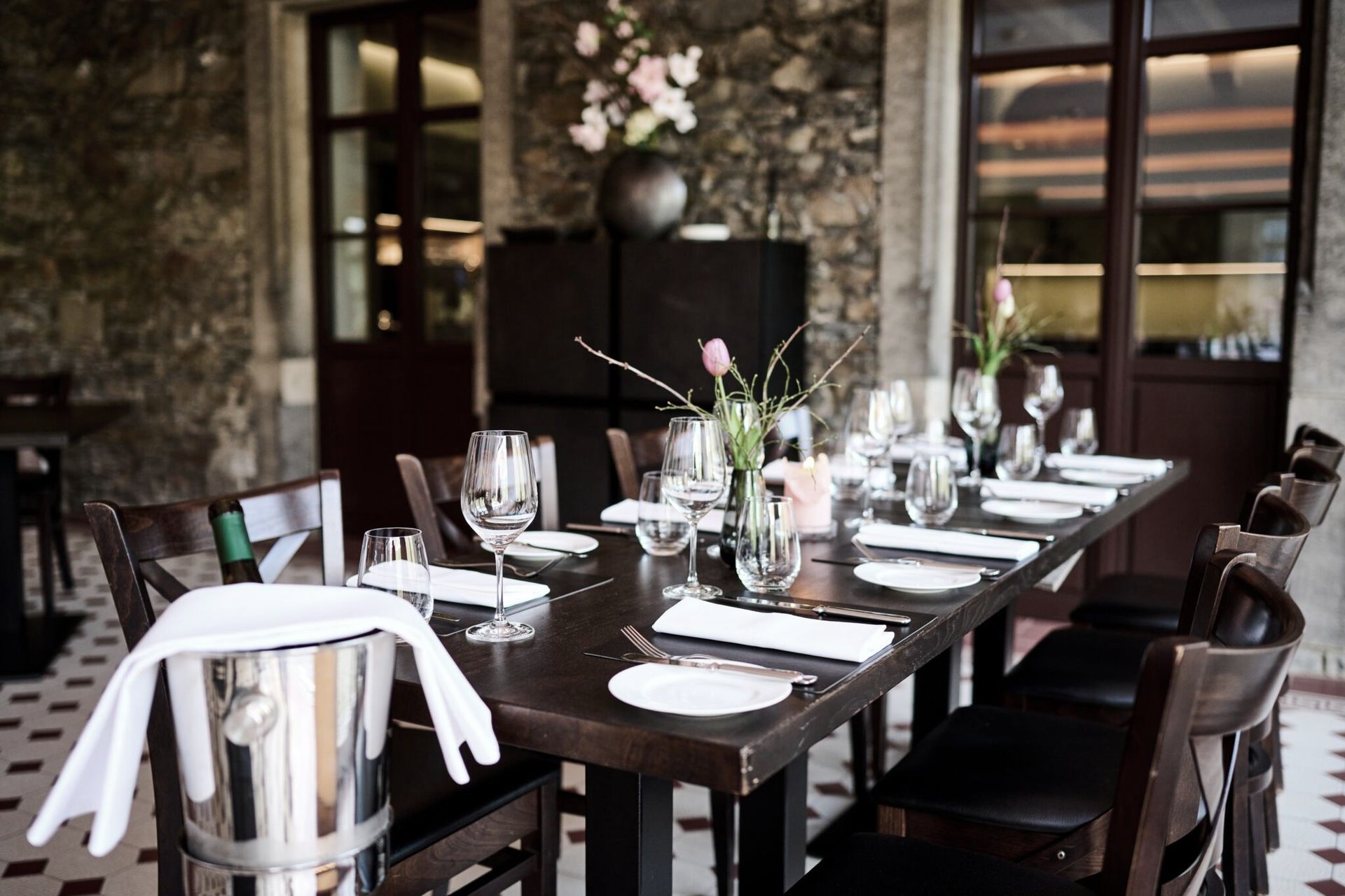 In einem rustikalen Steinrestaurant ist ein langer Esstisch aus Holz für Gäste gedeckt mit Weingläsern, Tellern und einem Weinkühler.