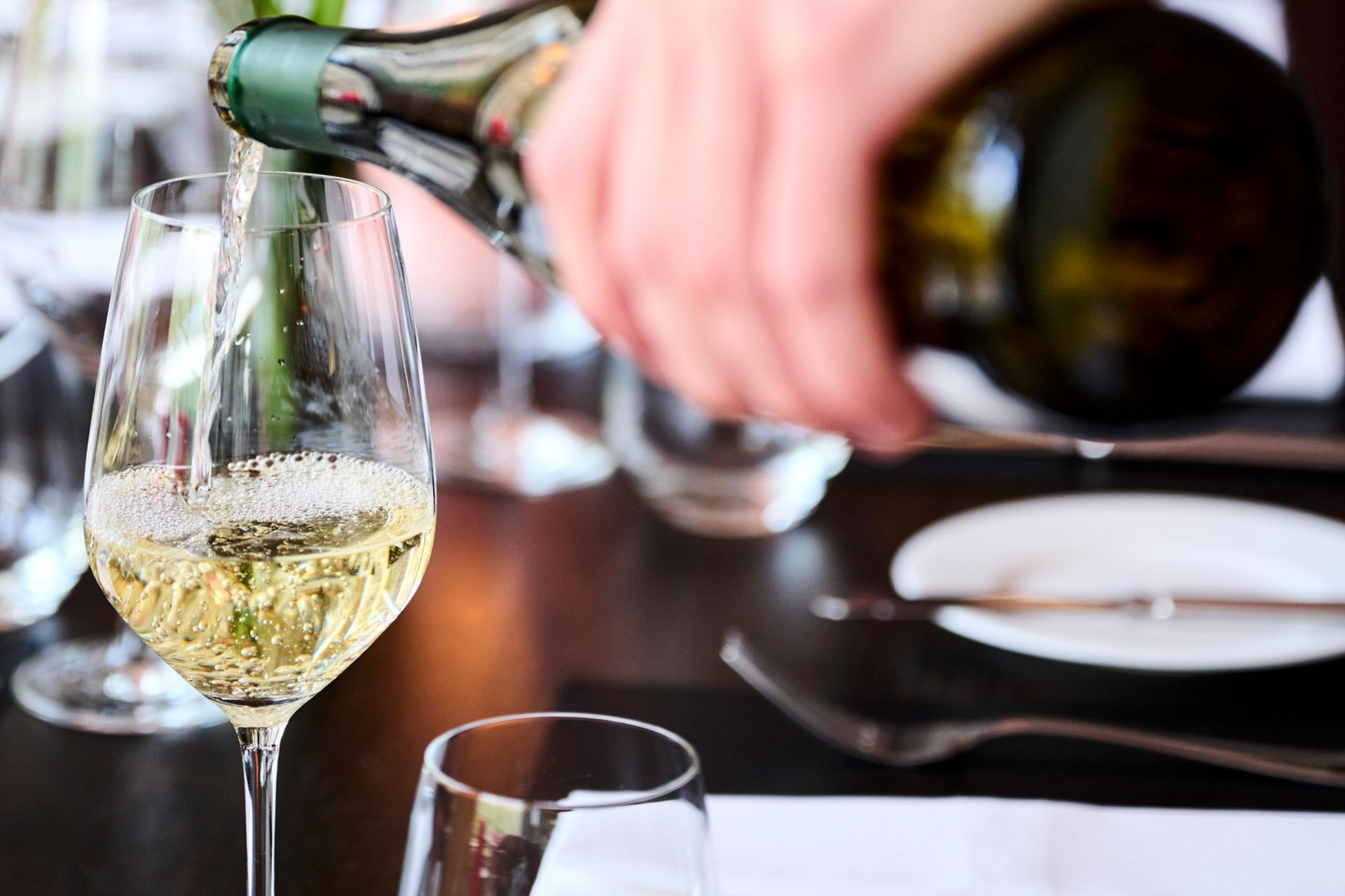 Eine Hand gießt Weißwein aus einer Flasche in ein Glas auf einem Tisch in gedeckter Essecke.