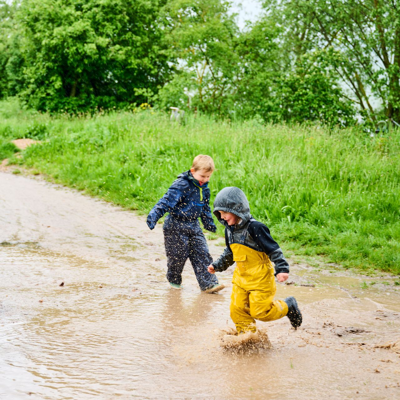 Zwei Kinder in Regenkleidung springen in eine schlammige Pfütze auf einem Feldweg, der von üppigem Grün umgeben ist.
