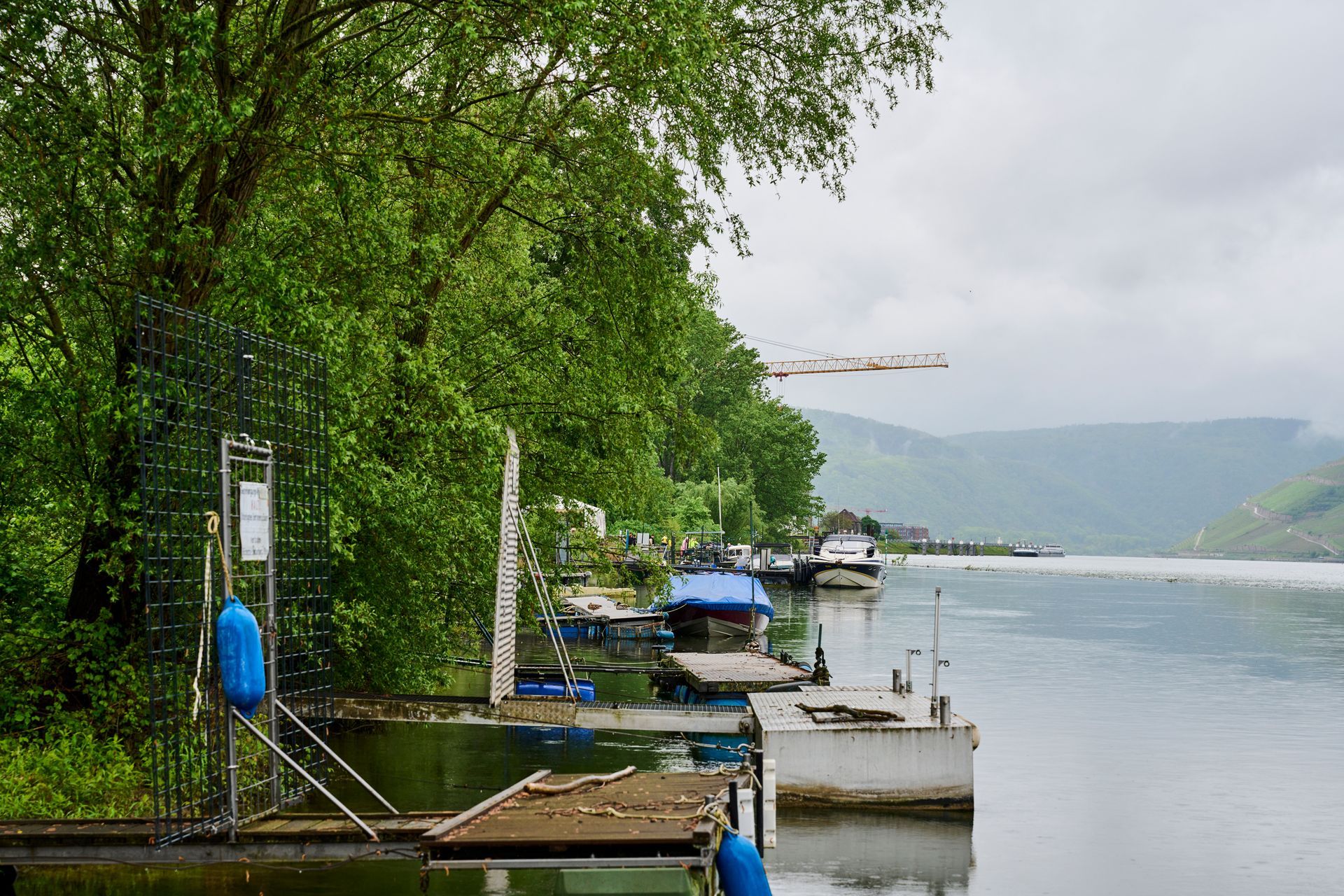 Ein üppig grünes Flussufer mit hölzernen Stegen und kleinen Booten unter bedecktem Himmel, im Hintergrund Berge.