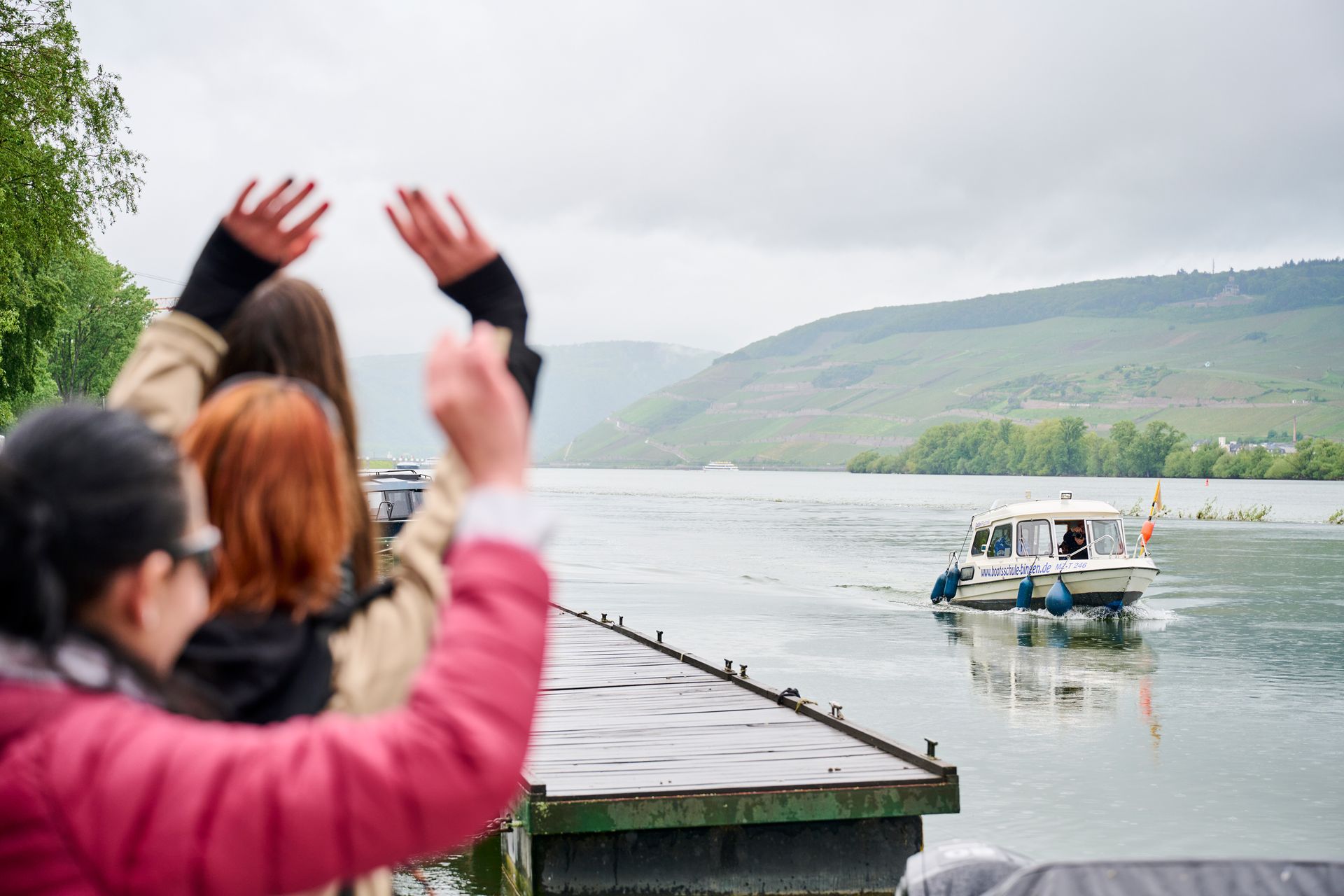 Drei Personen winken von hinten einem weißen Motorboot auf einem Fluss in der Nähe eines Docks und bewaldeter Hügel zu.