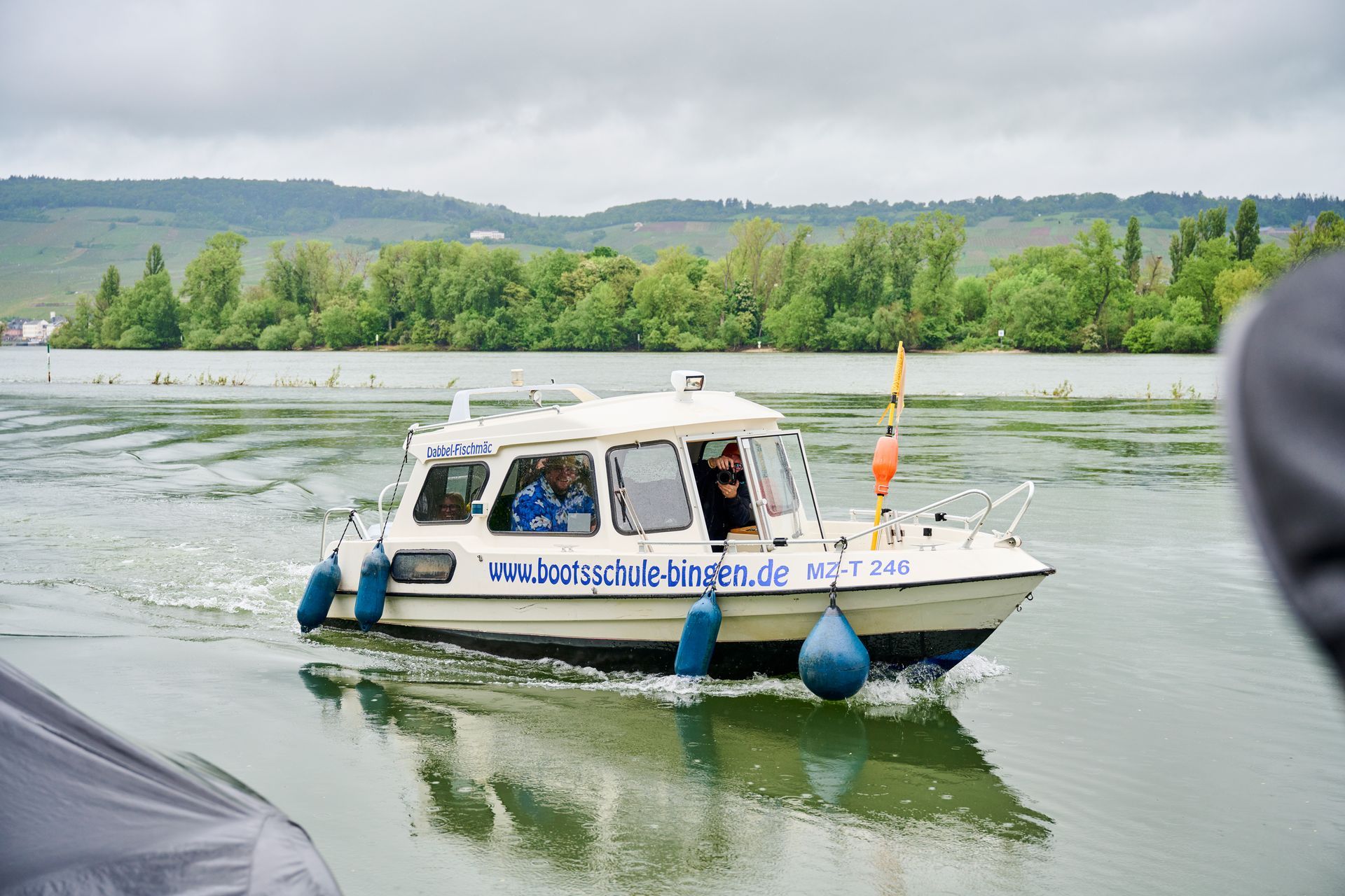 Ein weißes Motorboot mit blauen Fendern gleitet unter bedecktem Himmel auf einem ruhigen, von Bäumen gesäumten Fluss.
