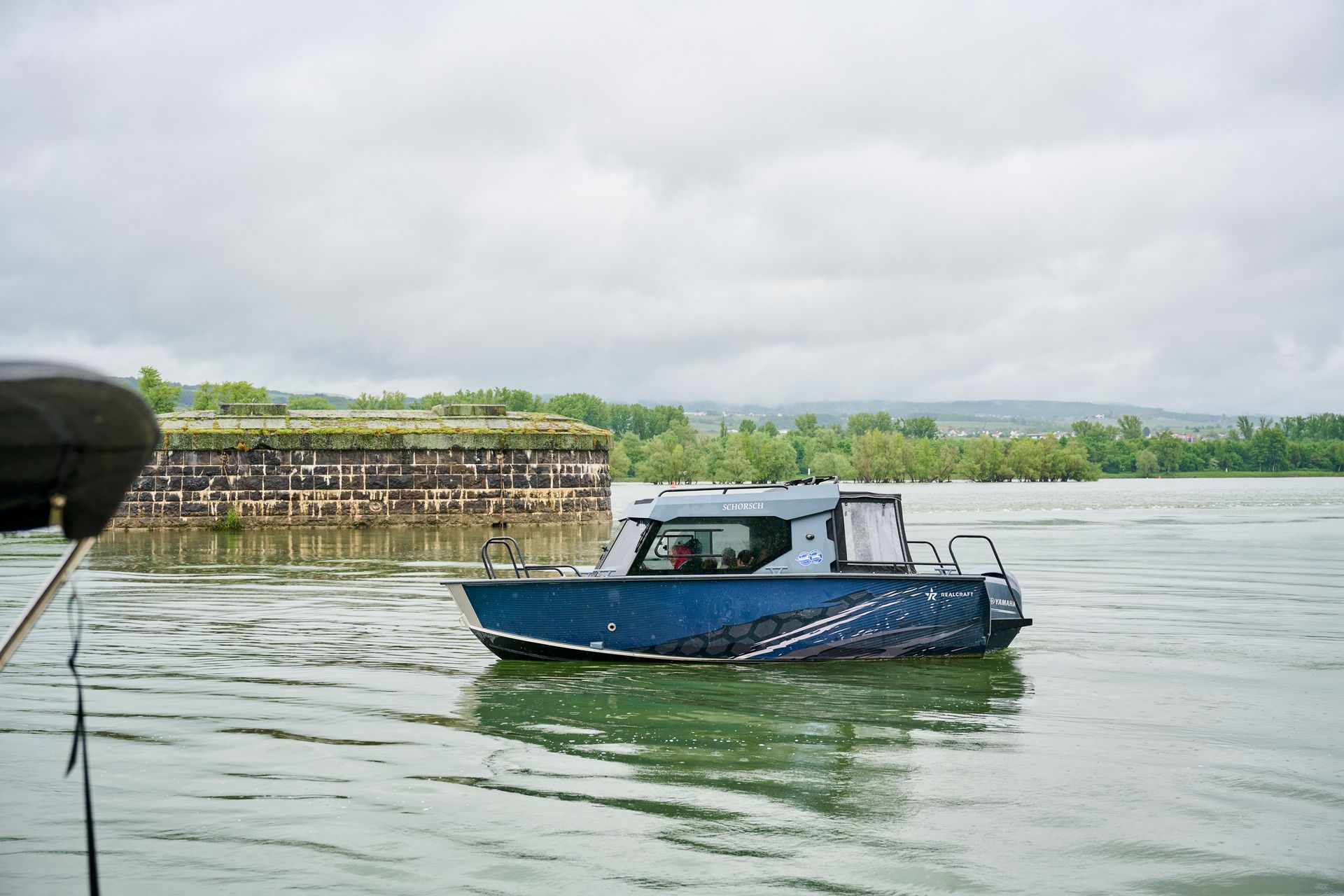Ein blaues Boot fährt auf ruhigem Wasser an einer steinernen Festungsanlage unter bewölktem Himmel vorbei.