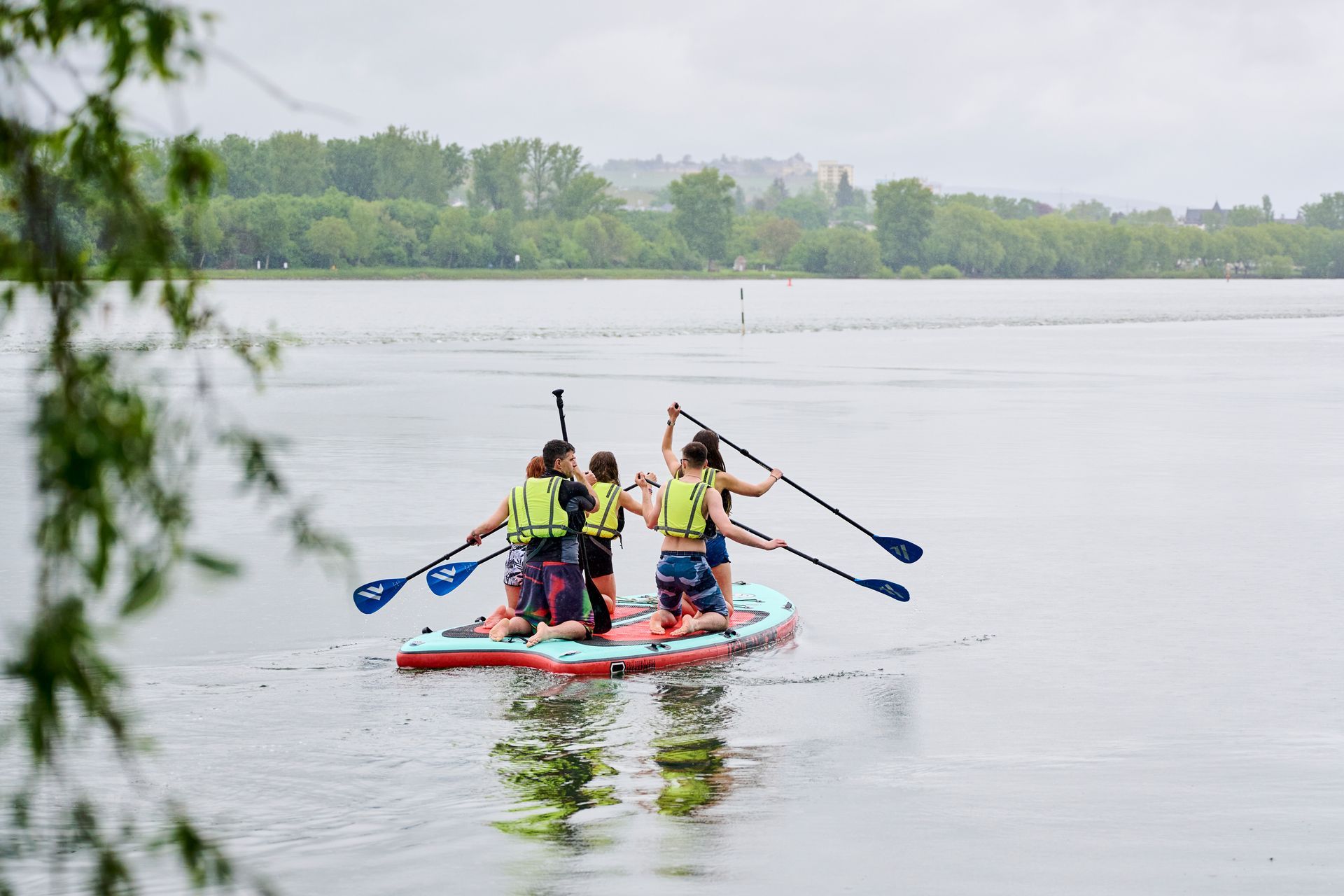 Four people wearing life jackets are kneeling on a large stand-up paddleboard on a lake and propelling themselves across the water with paddles.