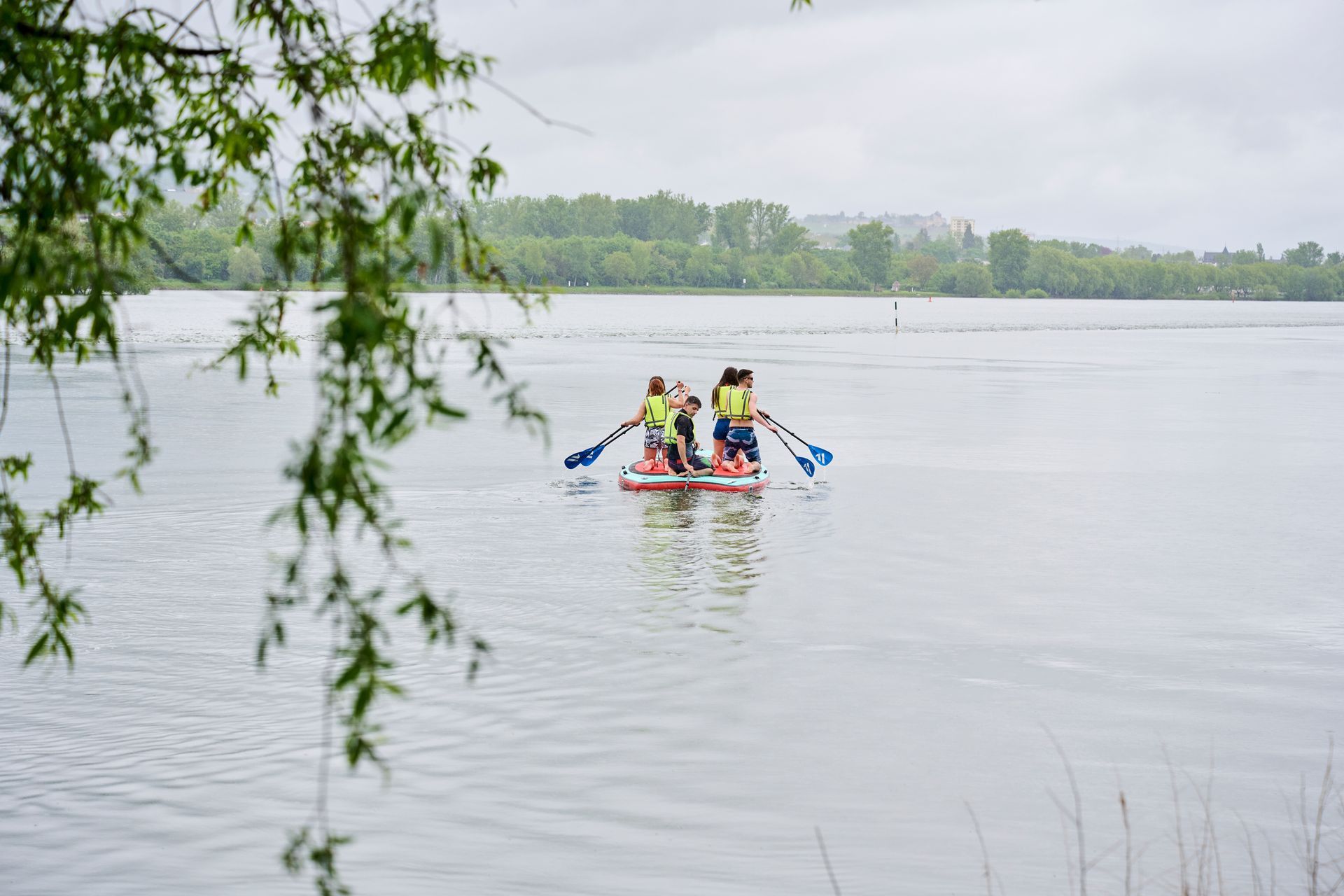 Vier Personen in Schwimmwesten paddeln an einem bewölkten Tag mit einem Schlauchboot über einen ruhigen, grauen See.