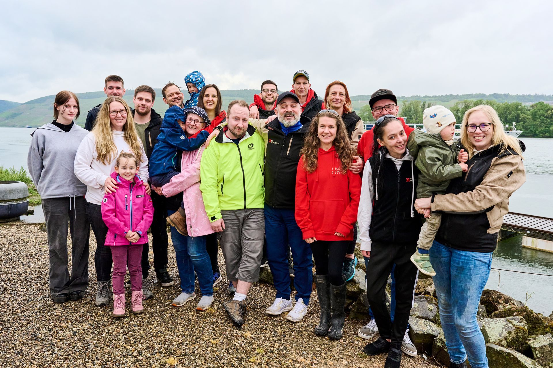 Eine große Gruppe posiert für ein Foto an einem felsigen Seeufer unter bedecktem Himmel.