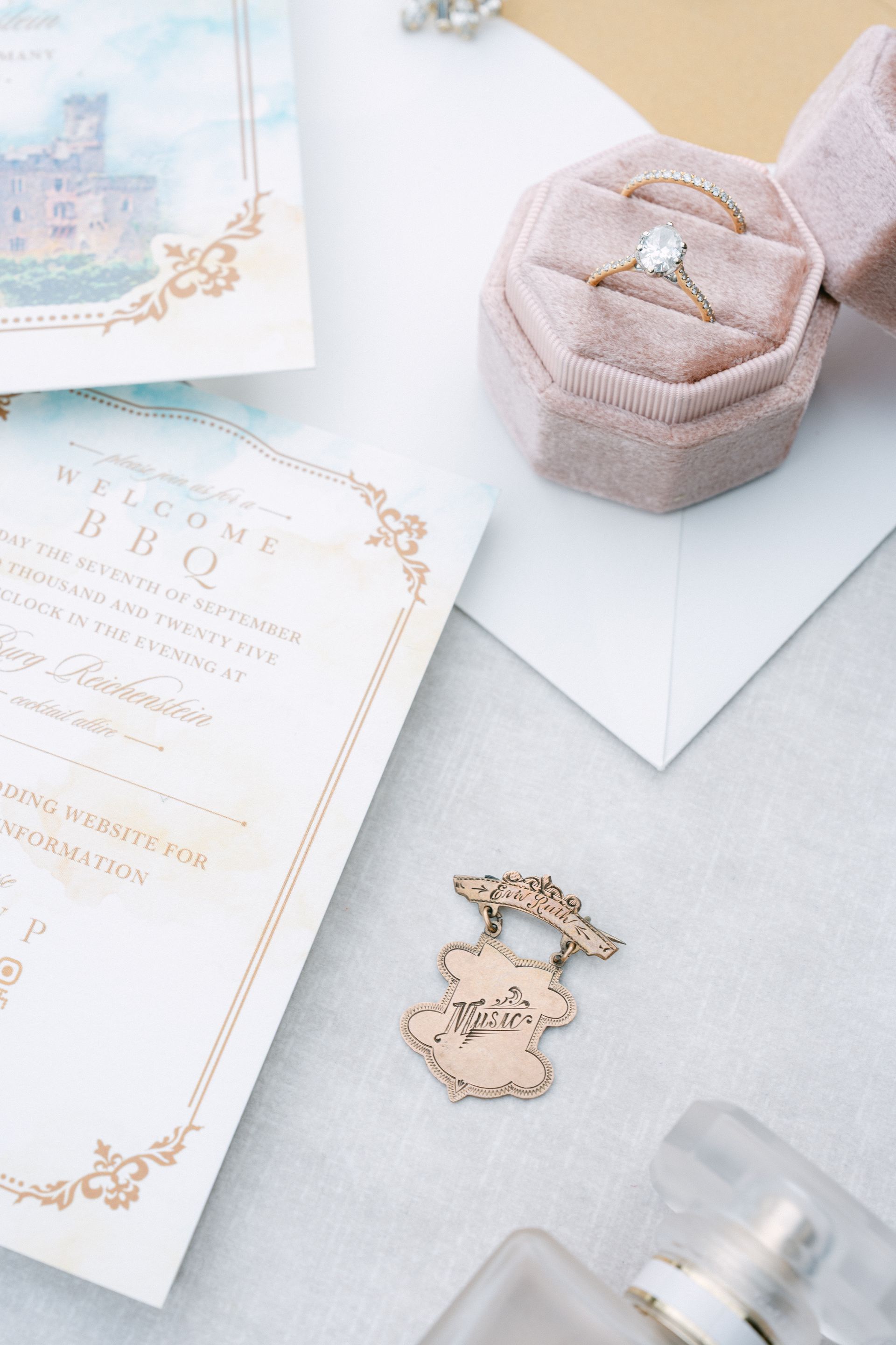 Ein flach liegendes Hochzeitsfoto mit einer Ringschachtel mit zwei Ringen, einer gedruckten Einladung und einem dekorativen Goldornament.