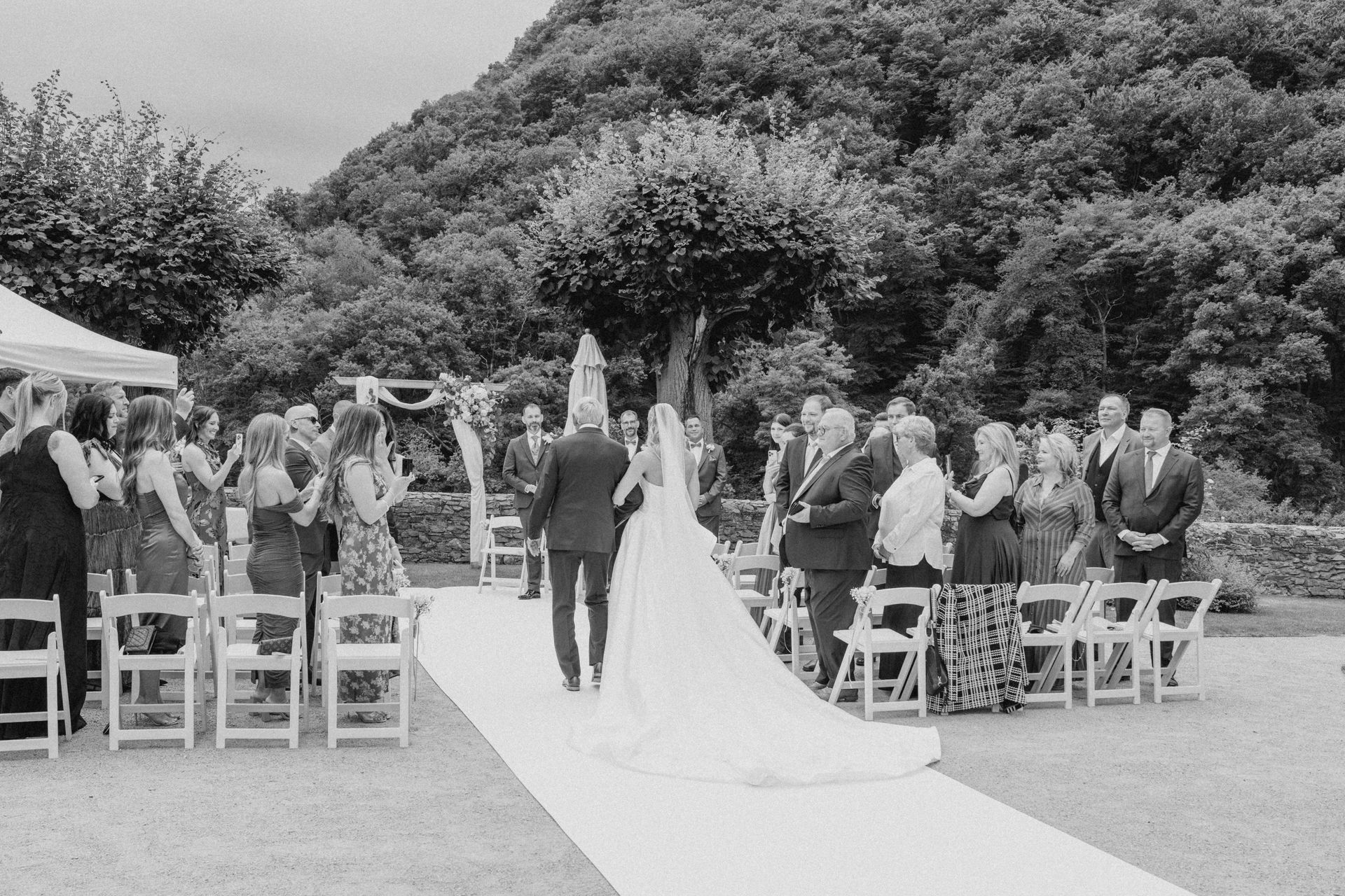 A couple walks down a white aisle outdoors in a garden setting, surrounded by seated guests and trees.