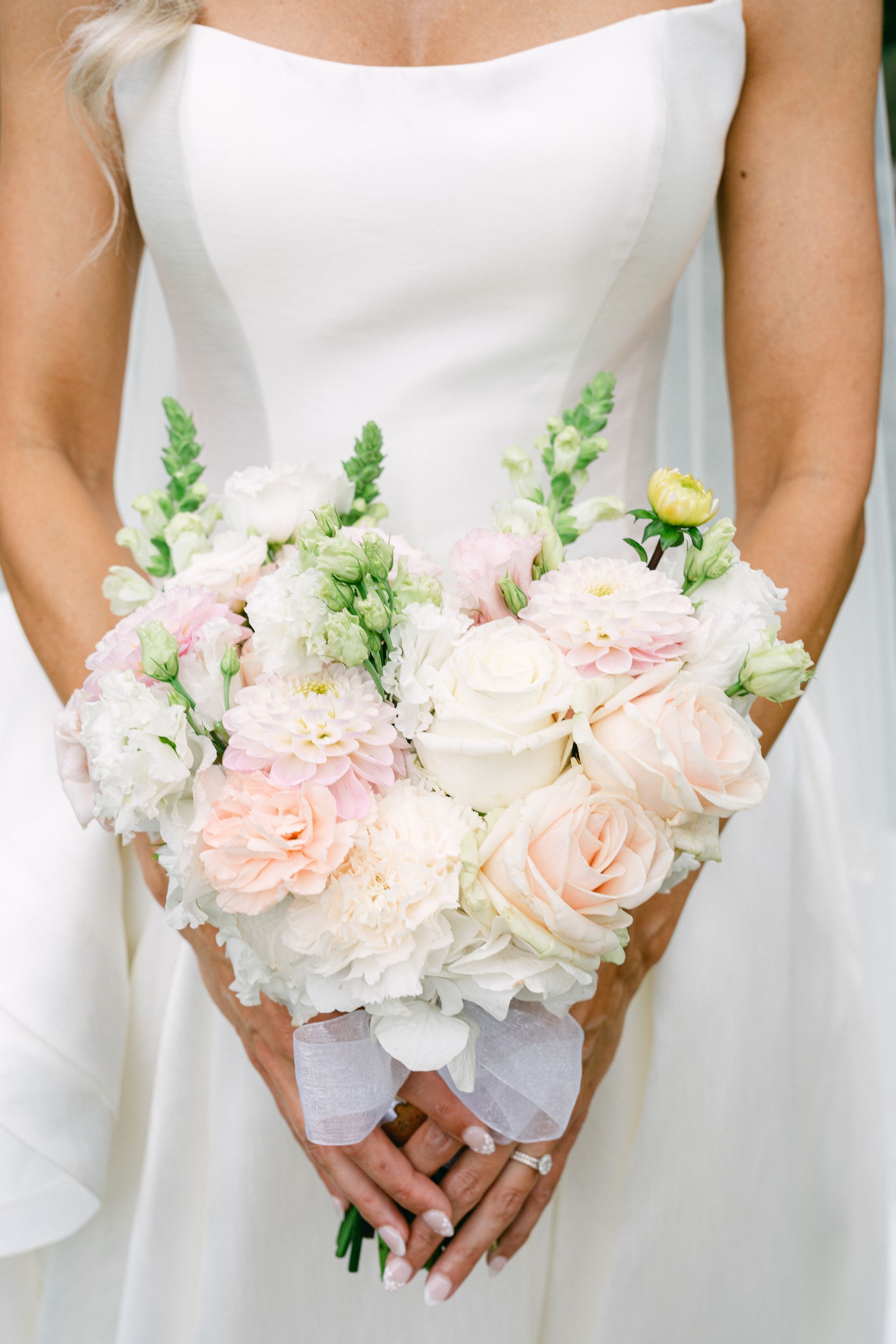 A person in a white dress is holding a bouquet of light pink and white roses, carnations, and snapdragons, tied together with a white ribbon.