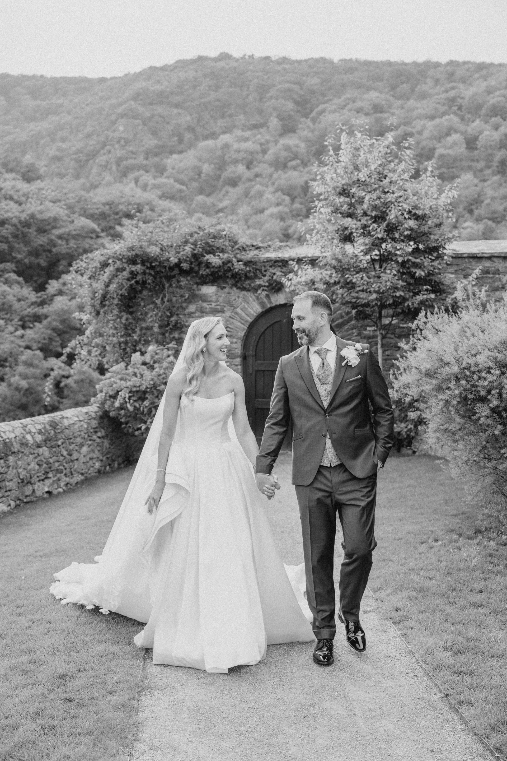 A black-and-white photo shows a bride and groom walking hand in hand along a stone path in front of a hill.