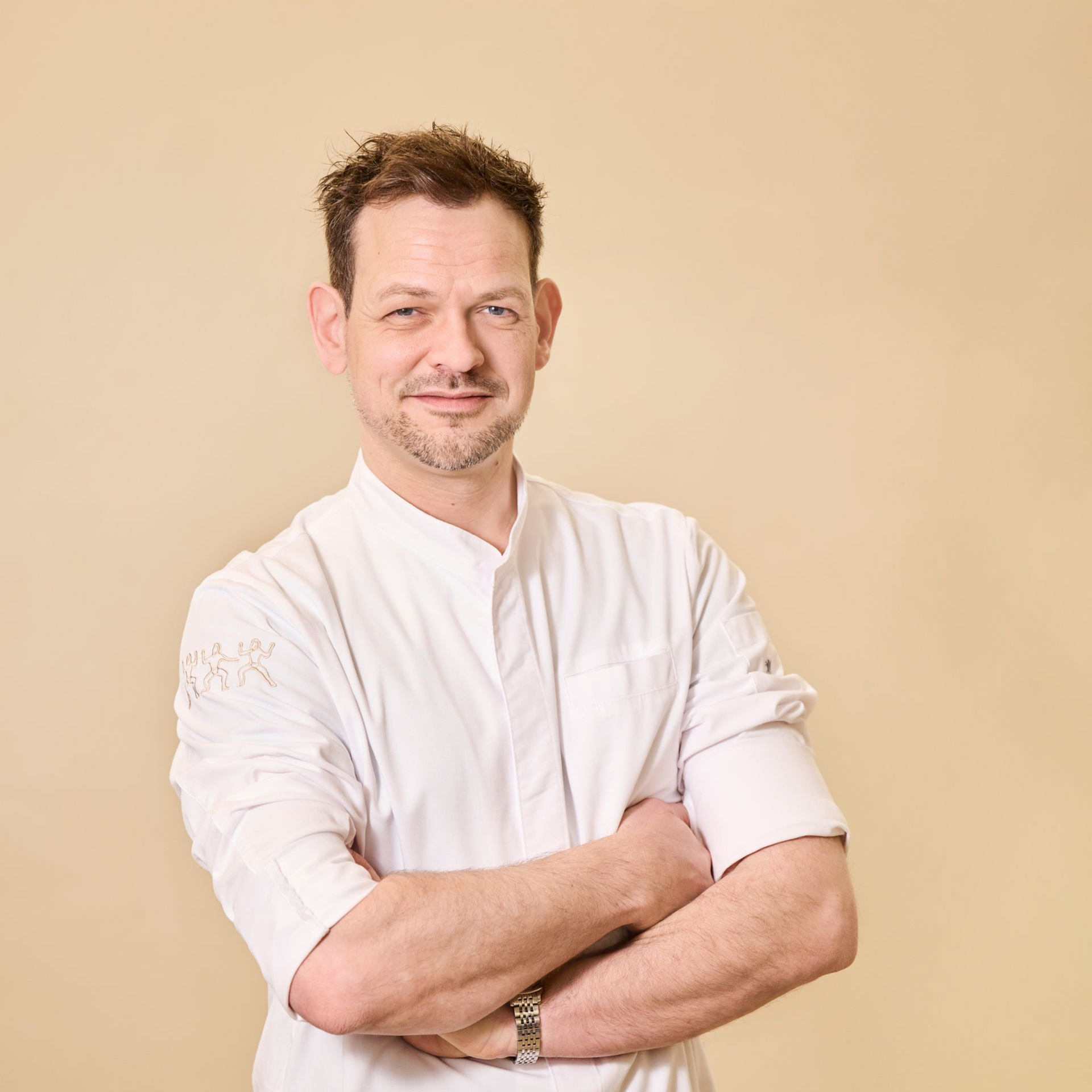 Eine Person in weißer Kochuniform steht mit verschränkten Armen vor einem beigen Hintergrund und blickt in die Kamera.