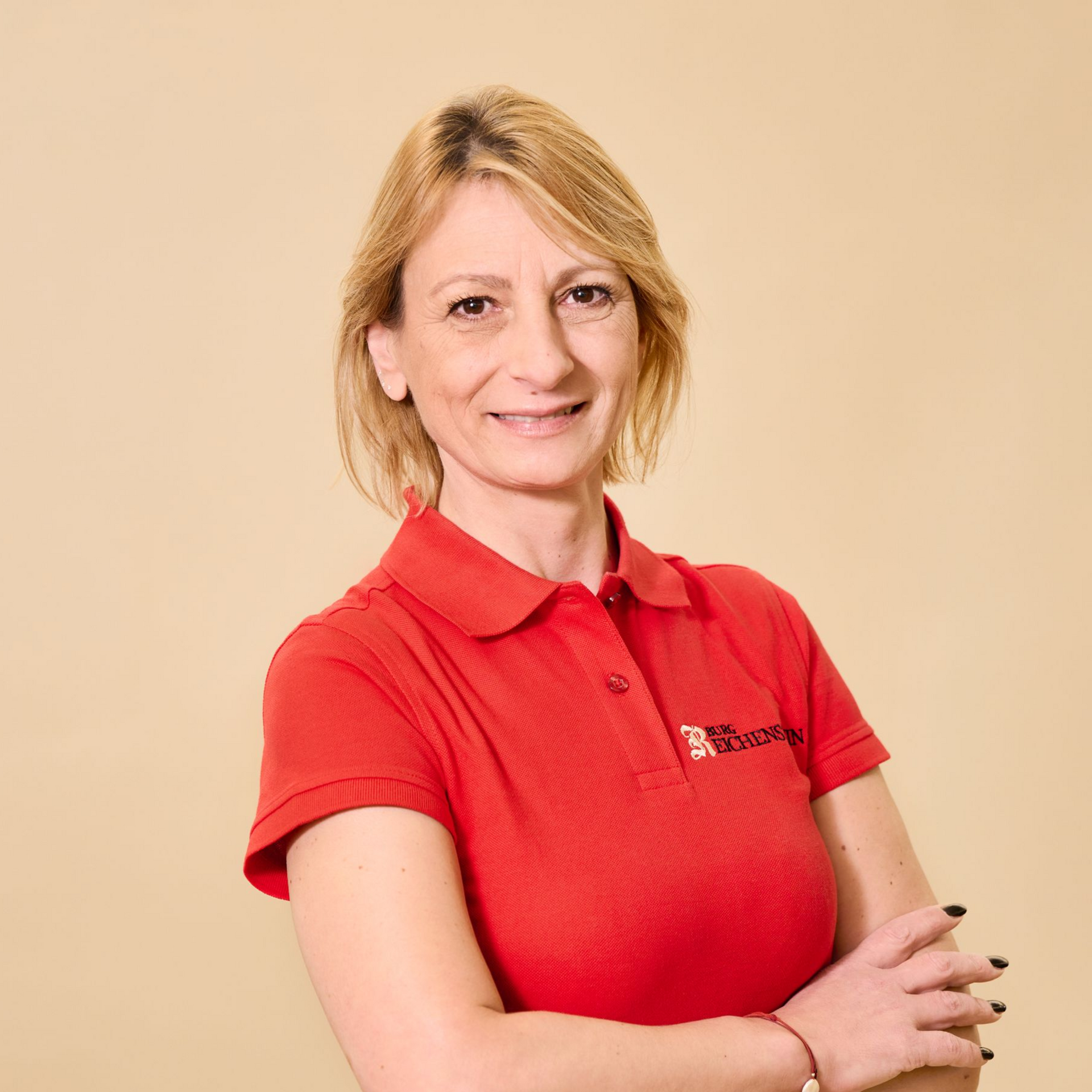 Eine Person mit blonden Haaren und einem roten Poloshirt steht mit verschränkten Armen vor einem einfarbigen, hellen Hintergrund.