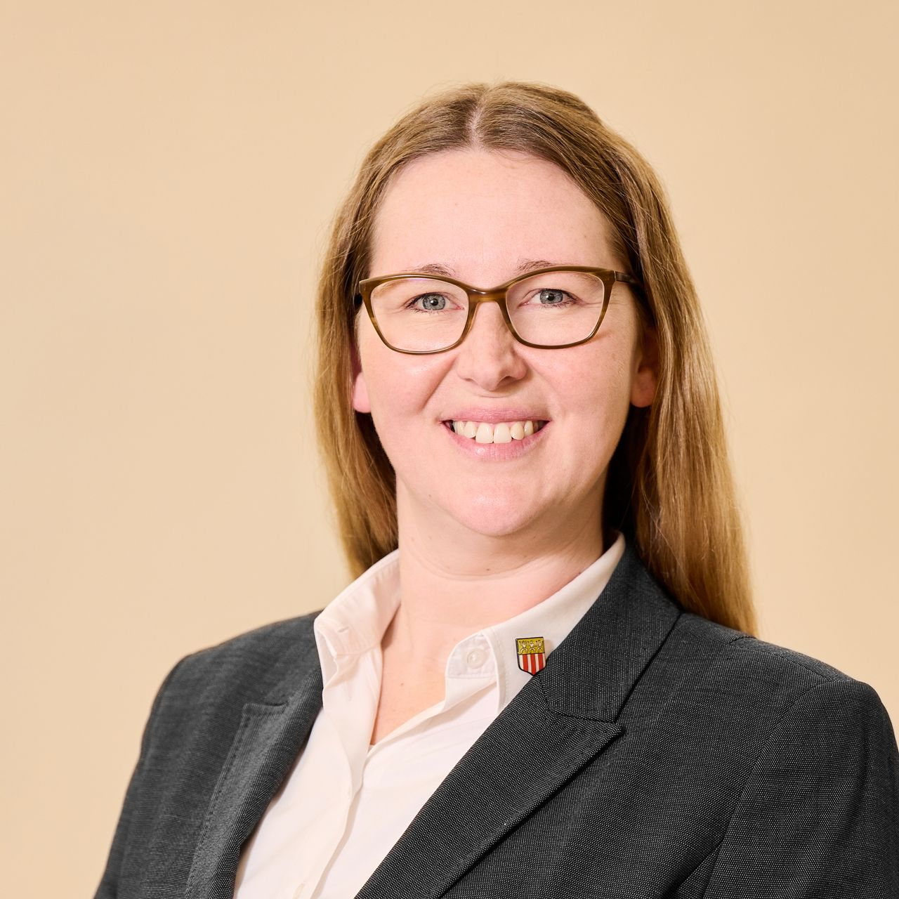 Eine lächelnde Person mit langen hellbraunen Haaren, die eine Brille, ein weißes Hemd mit Kragen und einen dunklen, strukturierten Blazer trägt.