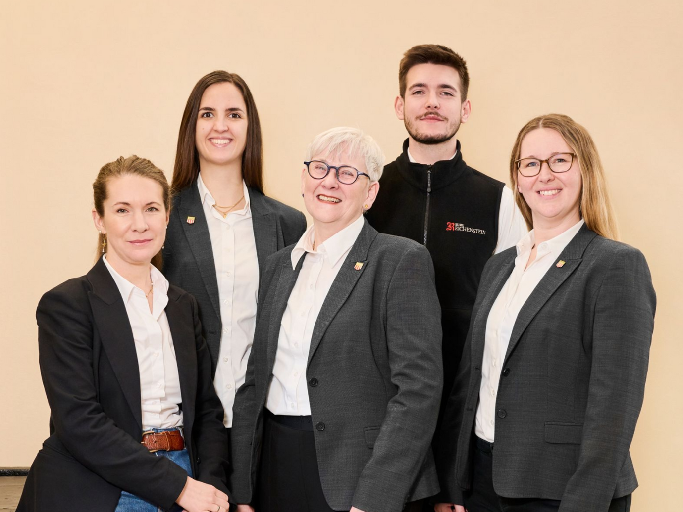 Eine Gruppe von fünf Personen in Business-Casual-Kleidung posiert vor einem schlichten, hellen Hintergrund.