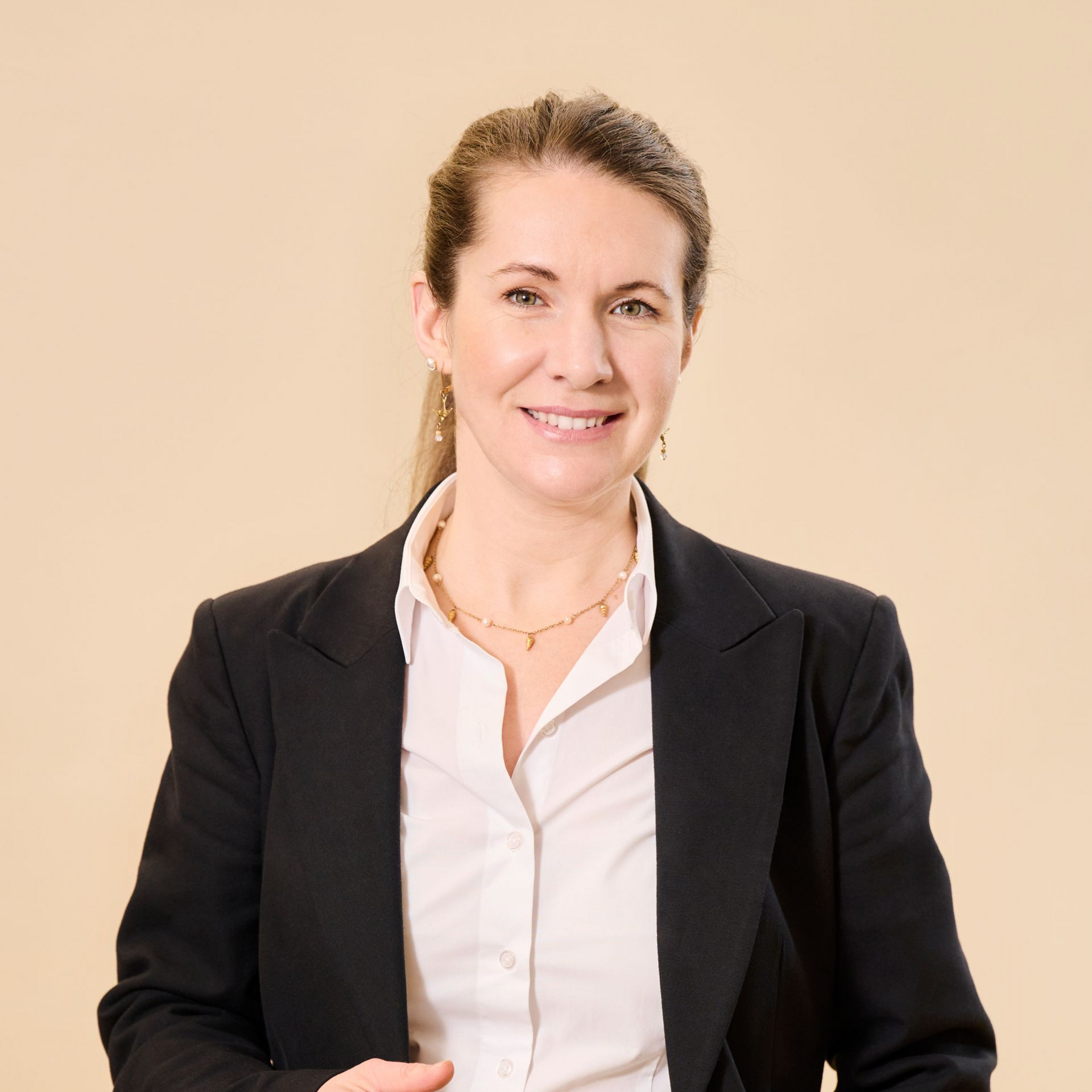 Ein professionelles Porträt einer Person mit langen braunen Haaren, die lächelt und ein weißes Hemd und einen schwarzen Blazer trägt, vor einer hellbraunen Wand.