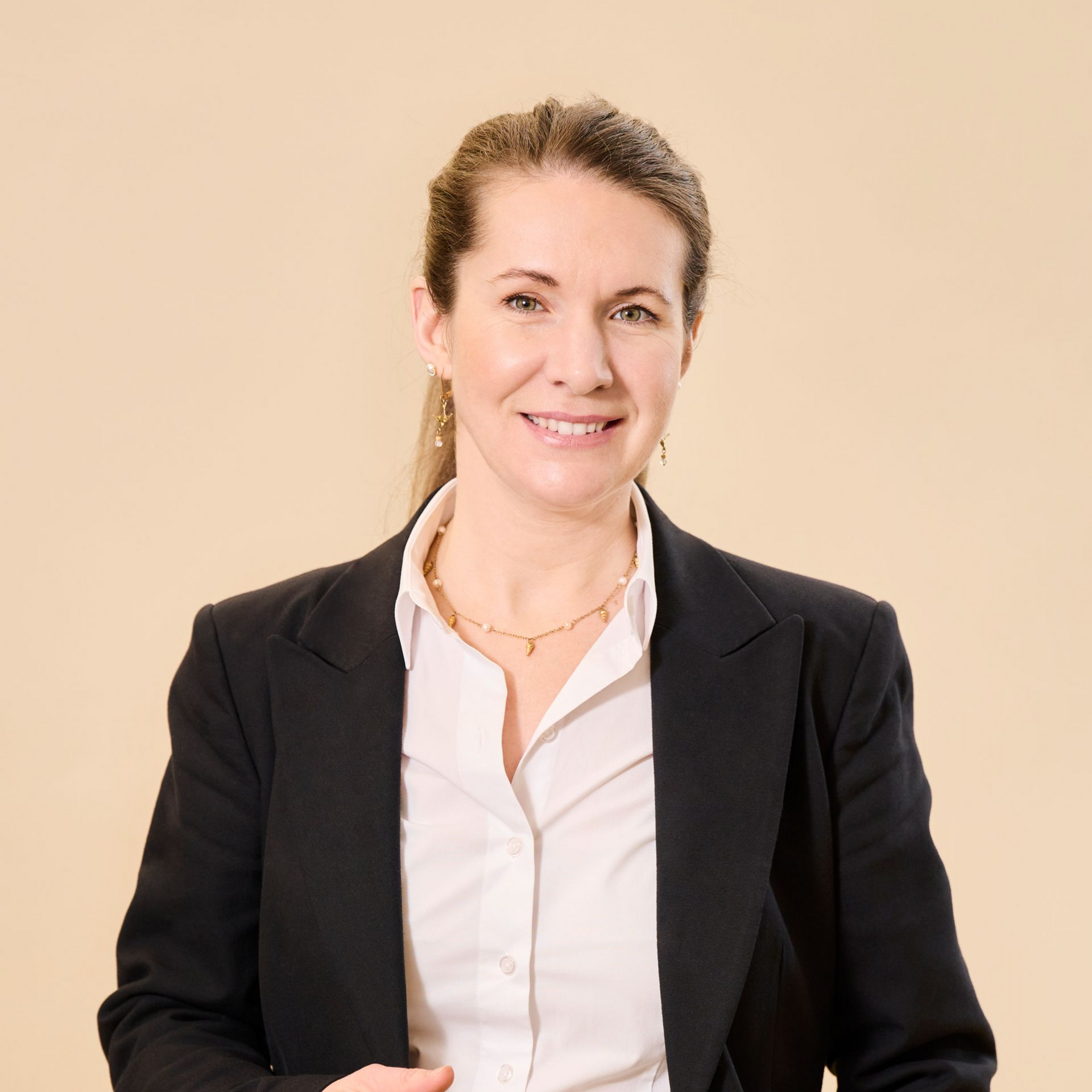 Eine lächelnde Person in einem dunklen Blazer und einem weißen Hemd mit Kragen vor einem einfarbig hellen Hintergrund.