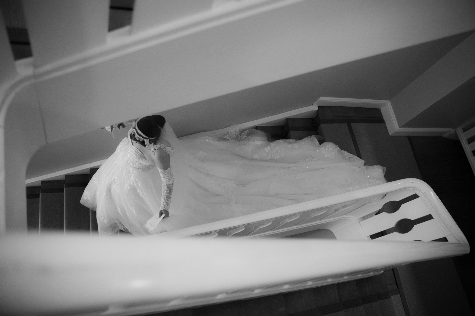 A person in a flowing white wedding dress is walking down a grand staircase; she is seen from above through the banister.