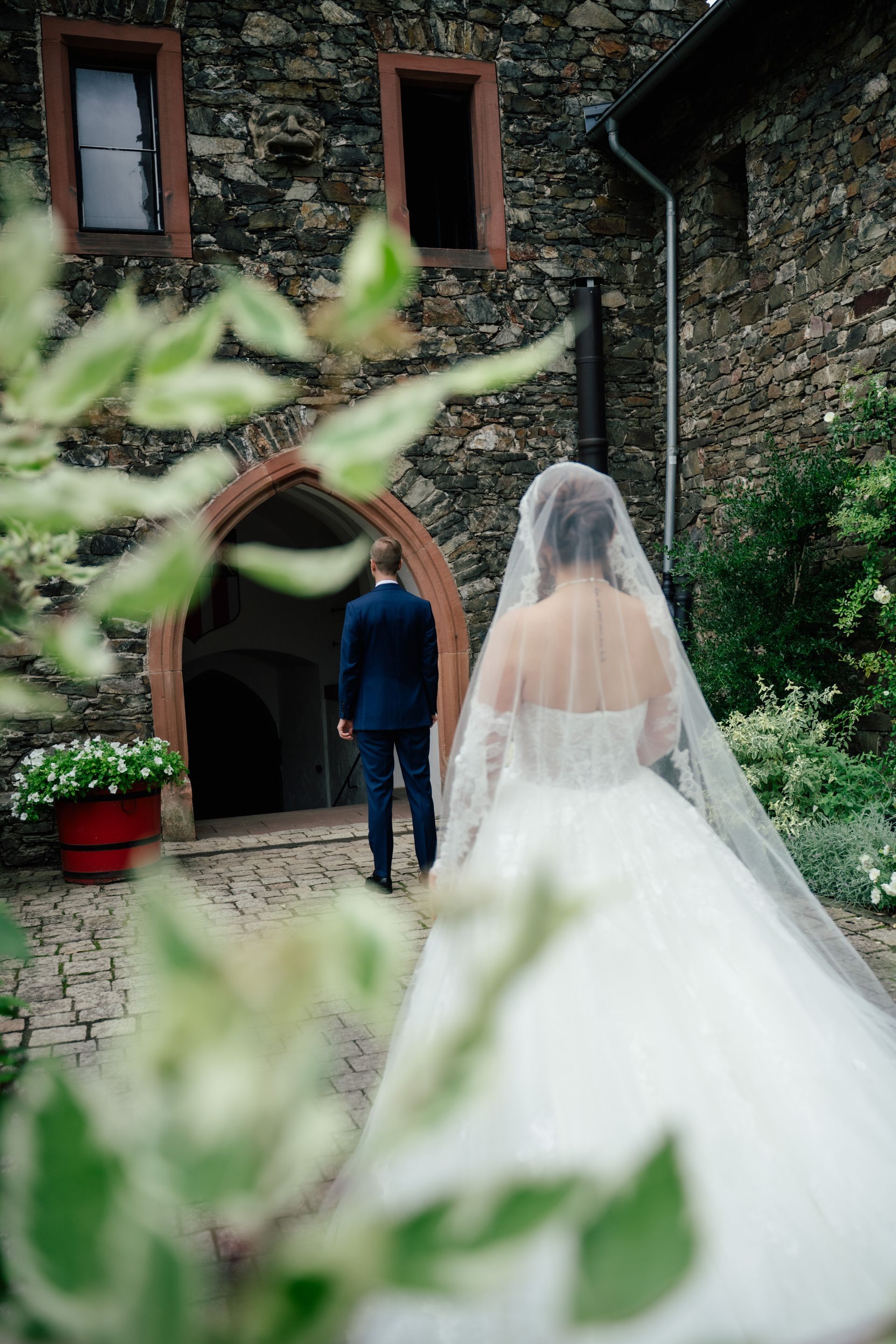 A bride in a white wedding dress approaches a groom standing in the doorway of a stone building, framed by blurred leaves.