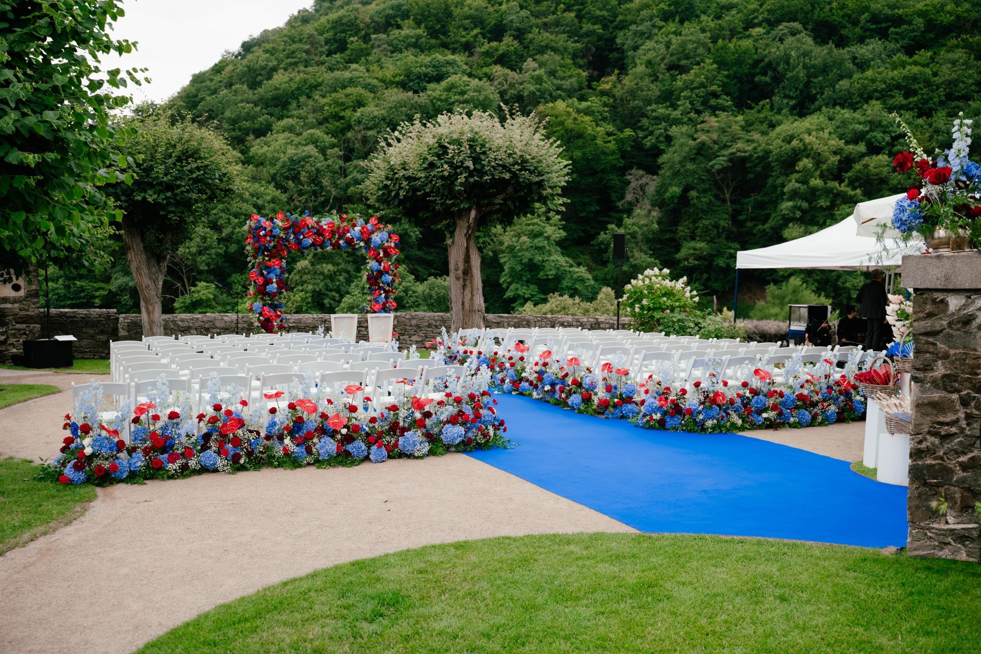 An outdoor wedding ceremony with white chairs, a blue carpet down the aisle, and red and blue floral arrangements.