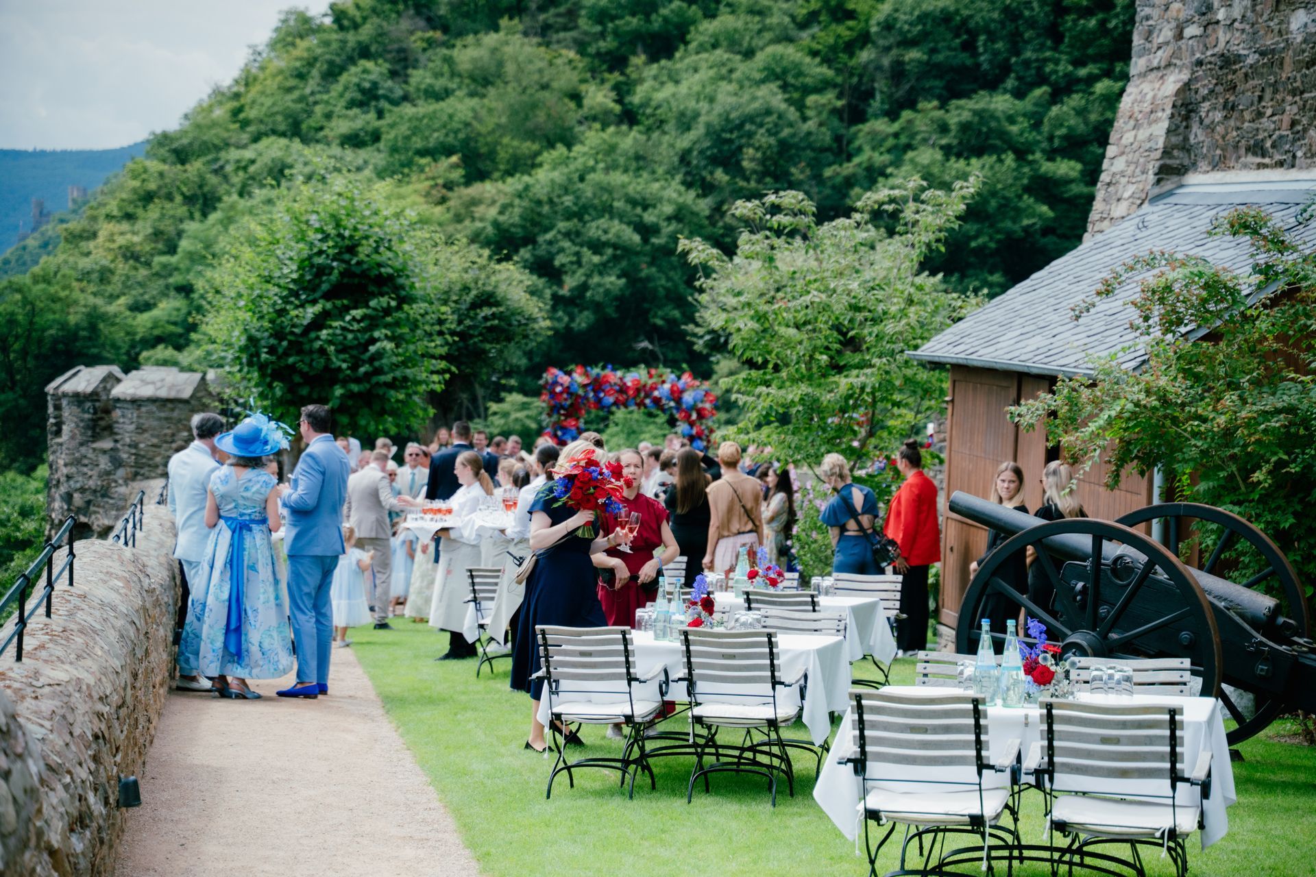 Guests at an outdoor wedding reception on a terraced lawn next to a stone castle, with round tables and a historic cannon.