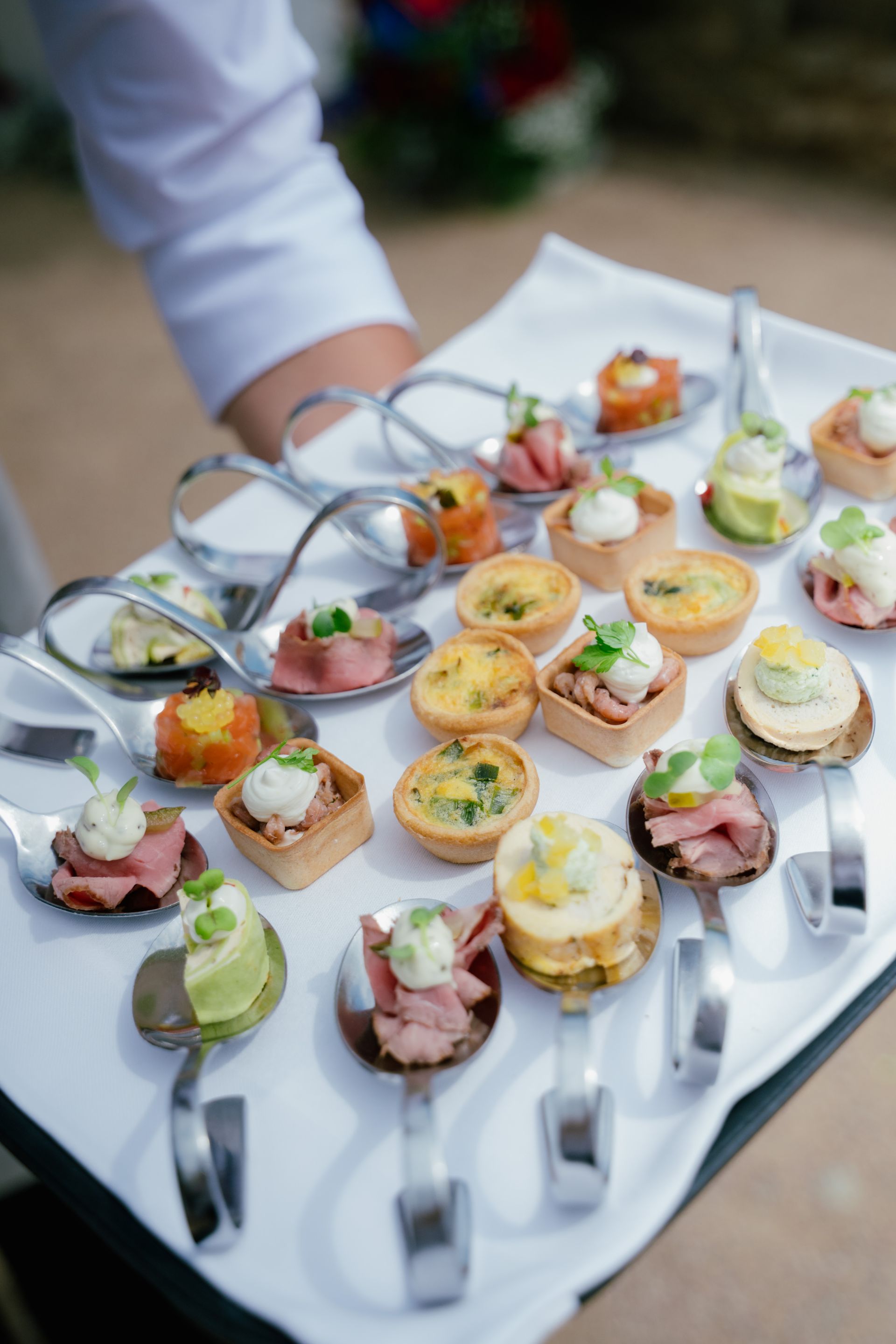 Ein Kellner trägt ein weißes Tablett, das mit verschiedenen Hors d'oeuvres auf Probierlöffeln und in kleinen, quadratischen Gebäckförmchen gefüllt ist.