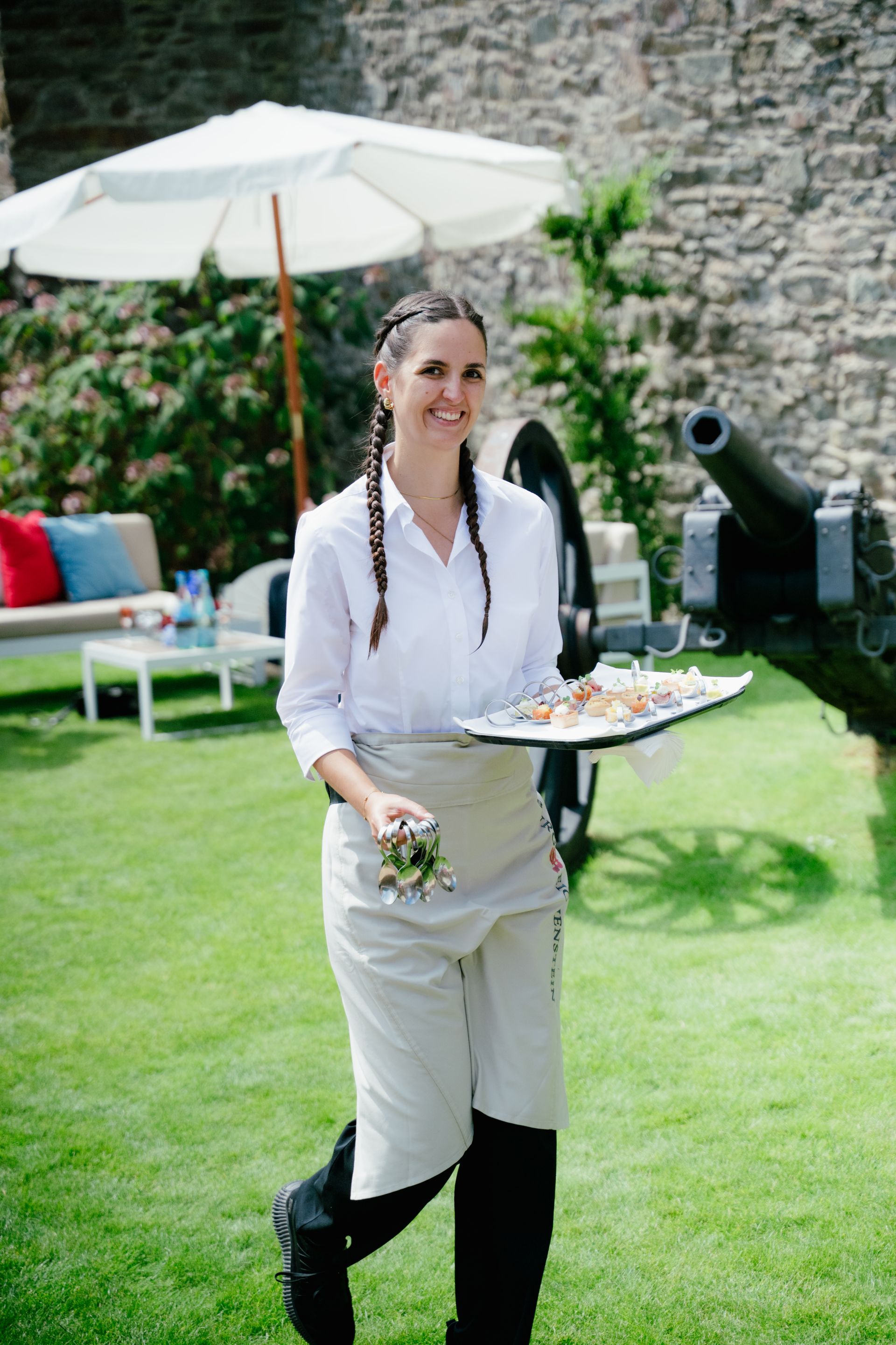 A smiling waitress in a white shirt and apron walks across a lawn near an outdoor cannon, carrying a tray full of appetizers.
