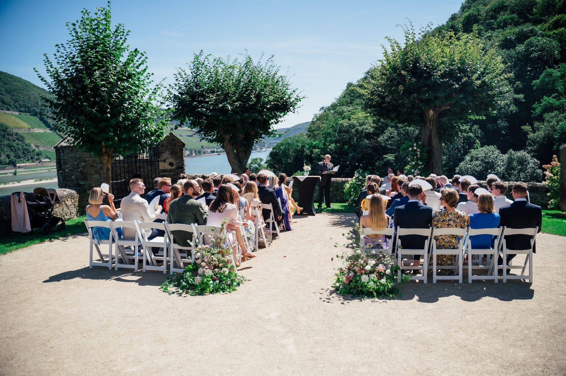 At an outdoor wedding ceremony overlooking a river and hills on a sunny day, the guests sit in rows of white chairs.