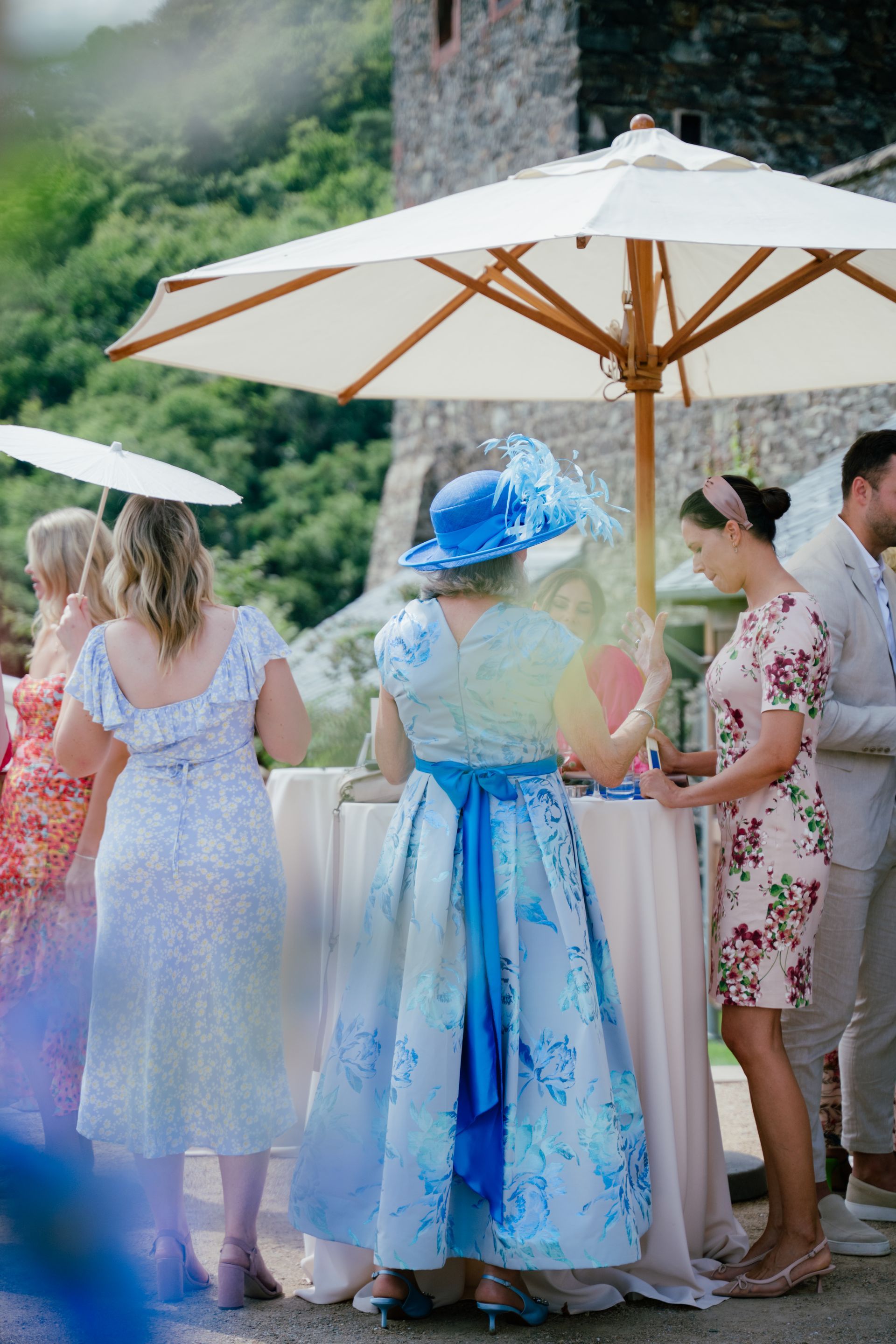 Gäste bei einer Veranstaltung im Freien unter einem weißen Sonnenschirm, im Vordergrund eine Person in einem blauen Blumenkleid und passendem Hut.