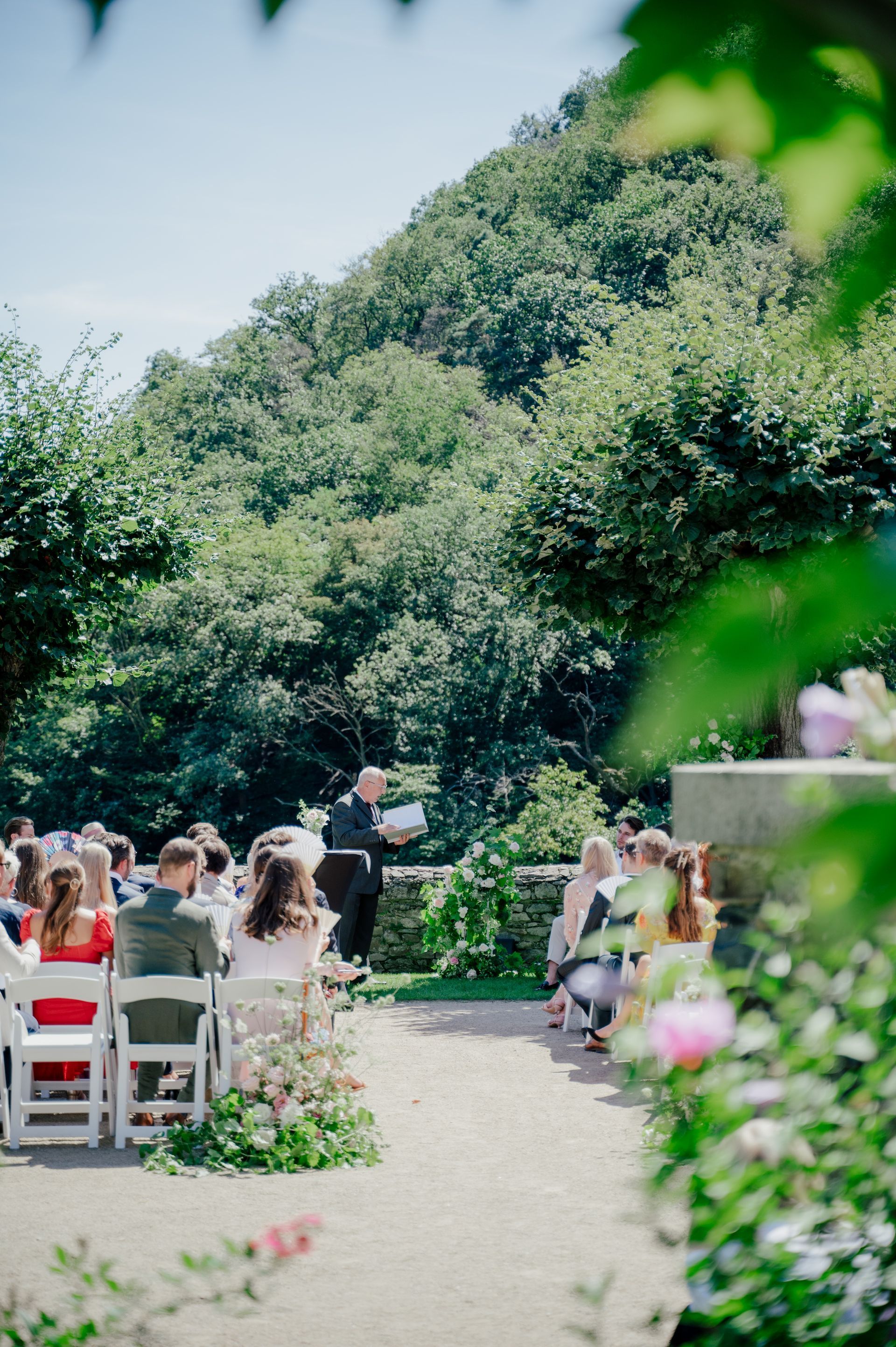 A wedding officiant presides over an outdoor wedding ceremony while the guests sit on folding chairs in front of a lush green hill.