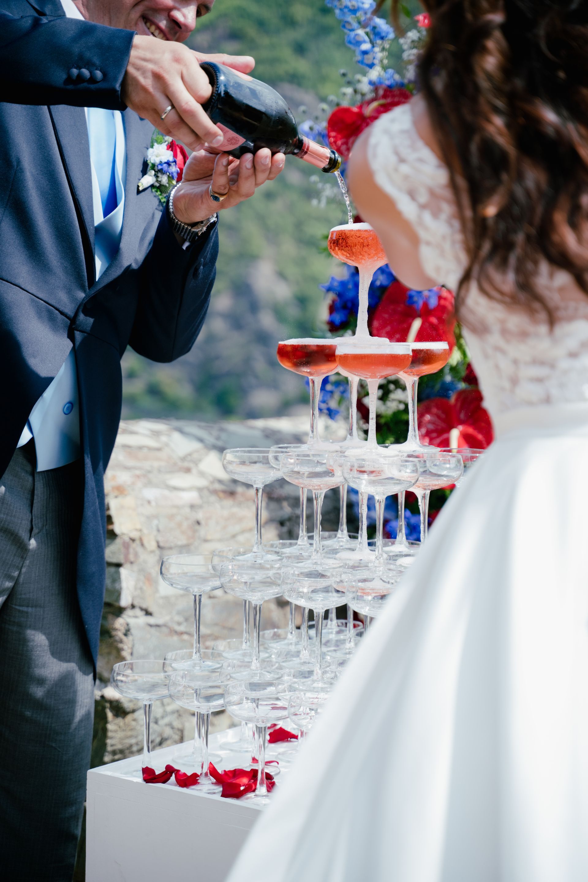 A man in a suit is pouring champagne into a multi-tiered tower of glasses, while a woman in a wedding dress stands next to him.