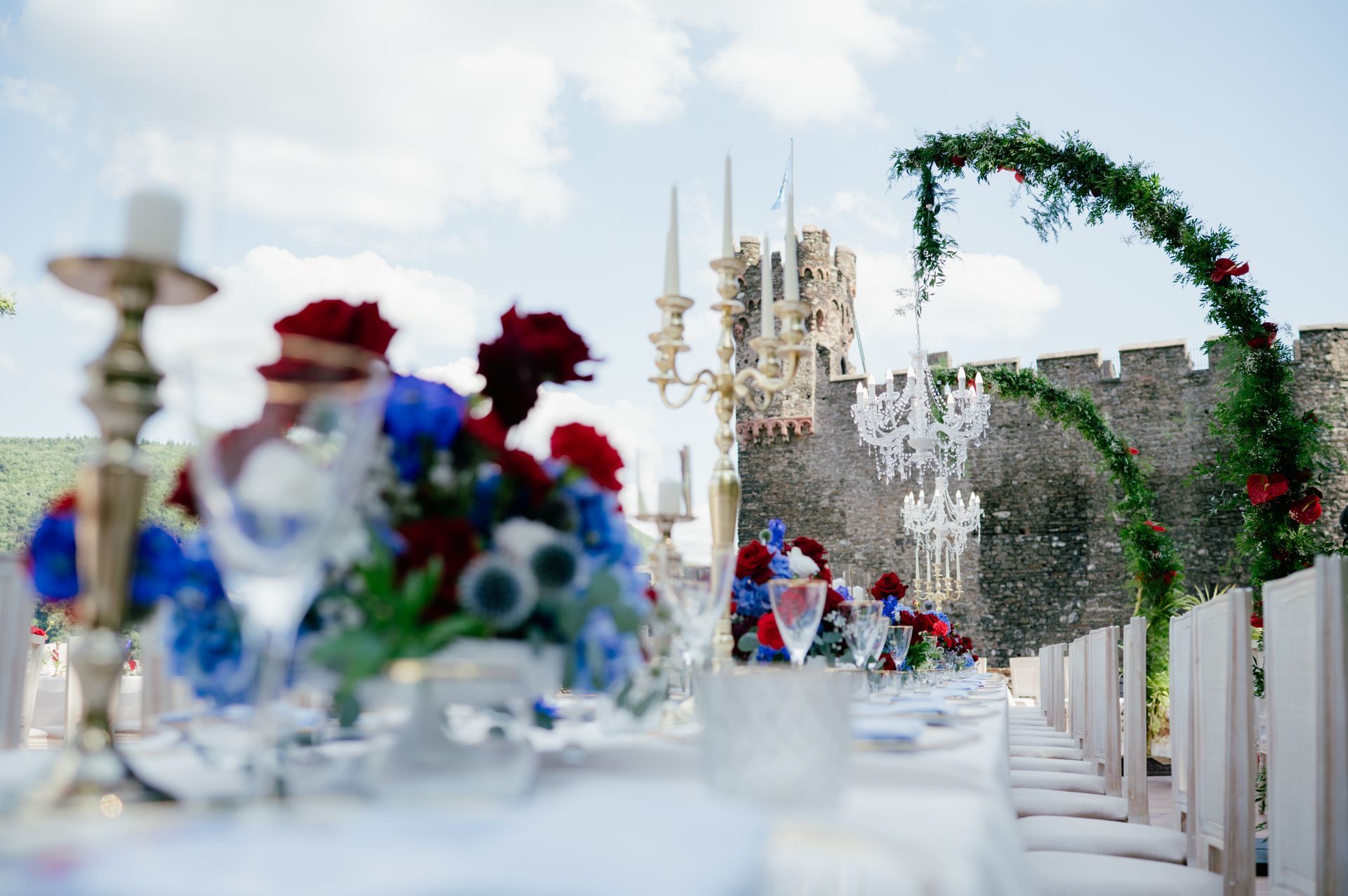 A long wedding table set outdoors, decorated with red and blue flowers and candelabras, with a stone castle in the background.