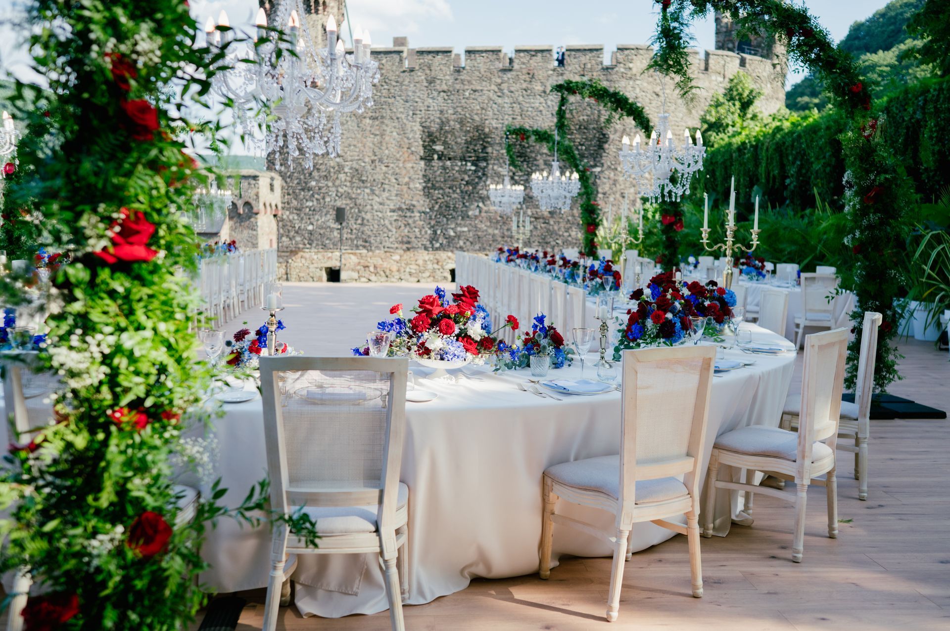 Eine lange, weiß gedeckte Hochzeitstafel mit roten und blauen Blumen, umgeben von weißen Stühlen in einem steinernen Schloss.