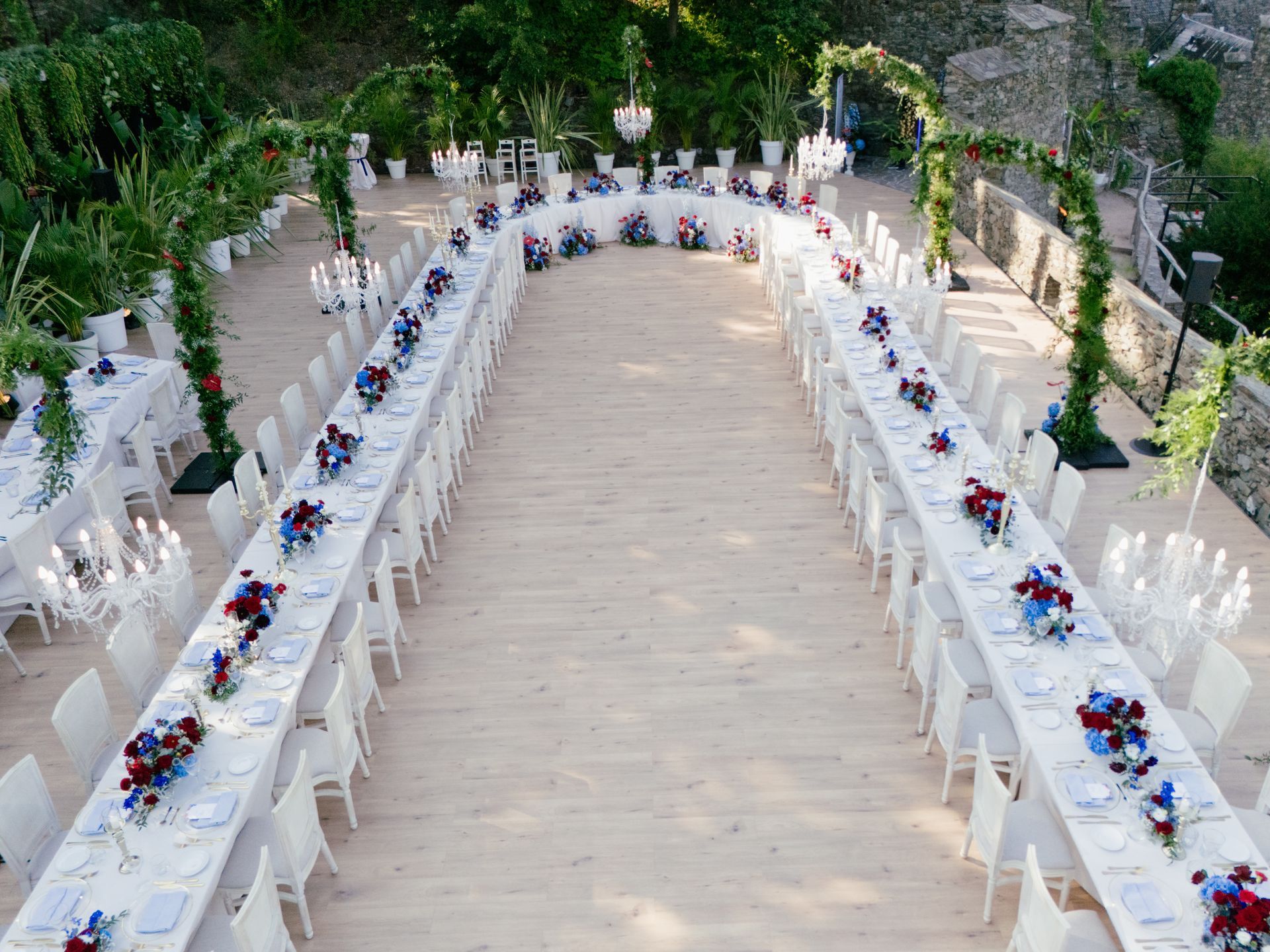 An outdoor U-shaped banquet table set up for a wedding, decorated with blue and red flowers and lit by string lights.