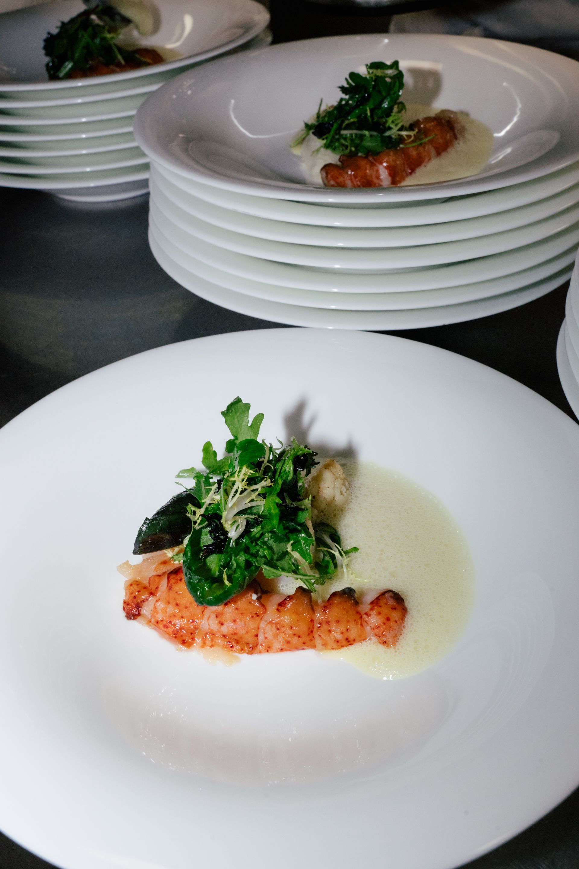 Ein auf einem Teller angerichteter Hummerschwanz mit frischem Gemüse und einer leichten Sauce, im Hintergrund Stapel sauberer weißer Teller.