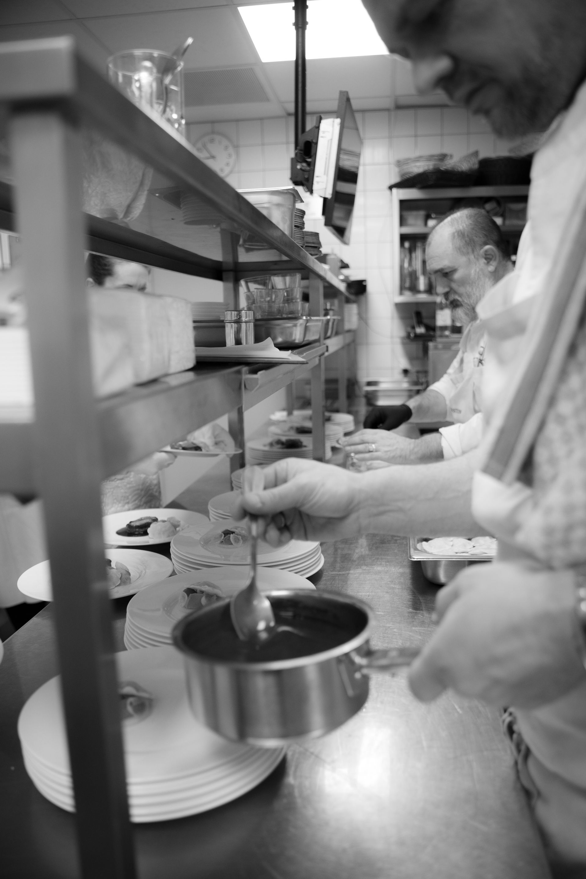 Menschen bereiten Essen in einer geschäftigen Küche zu; ein Topf und Küchenutensilien stehen auf der Arbeitsfläche.