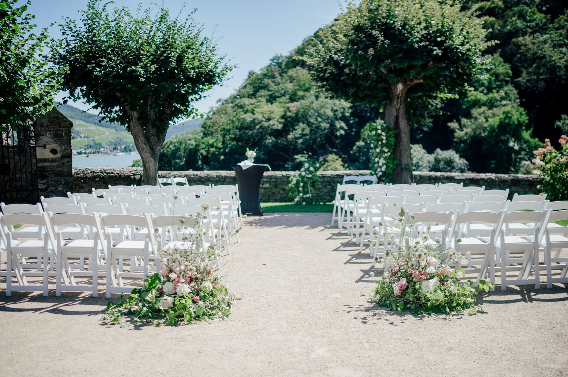 Aufbau für eine Hochzeitszeremonie im Freien mit Reihen weißer Stühle, die auf einem Kiesweg in der Nähe eines Flusses um ein zentrales Rednerpult angeordnet sind.