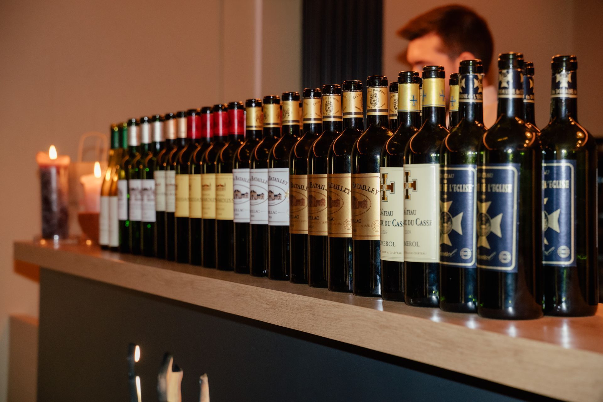 Auf einer Bartheke steht eine Reihe Weinflaschen, im Hintergrund sind eine verschwommene Person und zwei brennende Kerzen zu sehen.