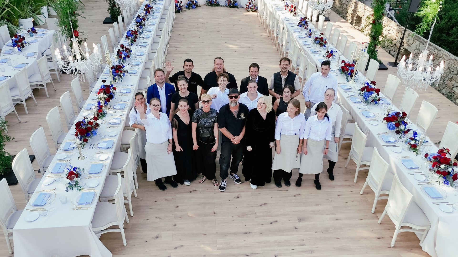 Eine Gruppe von Restaurantangestellten in Uniformen steht zwischen zwei langen, elegant gedeckten Esstischen in einem Außenbereich.