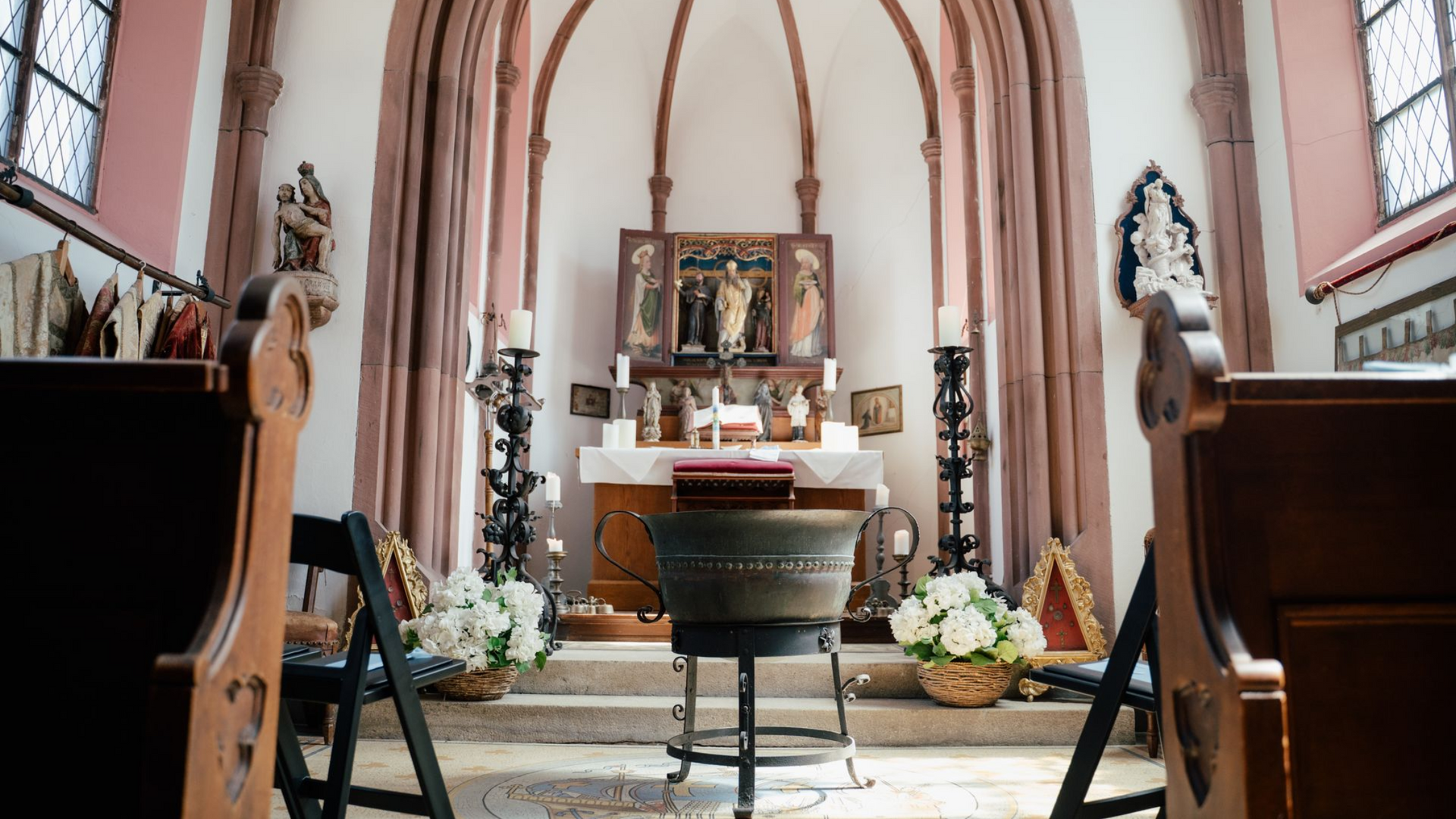 Der Altarraum einer Kirche mit einem zentralen Taufbecken aus Metall, Blumenarrangements und einem bemalten Holztriptychon.