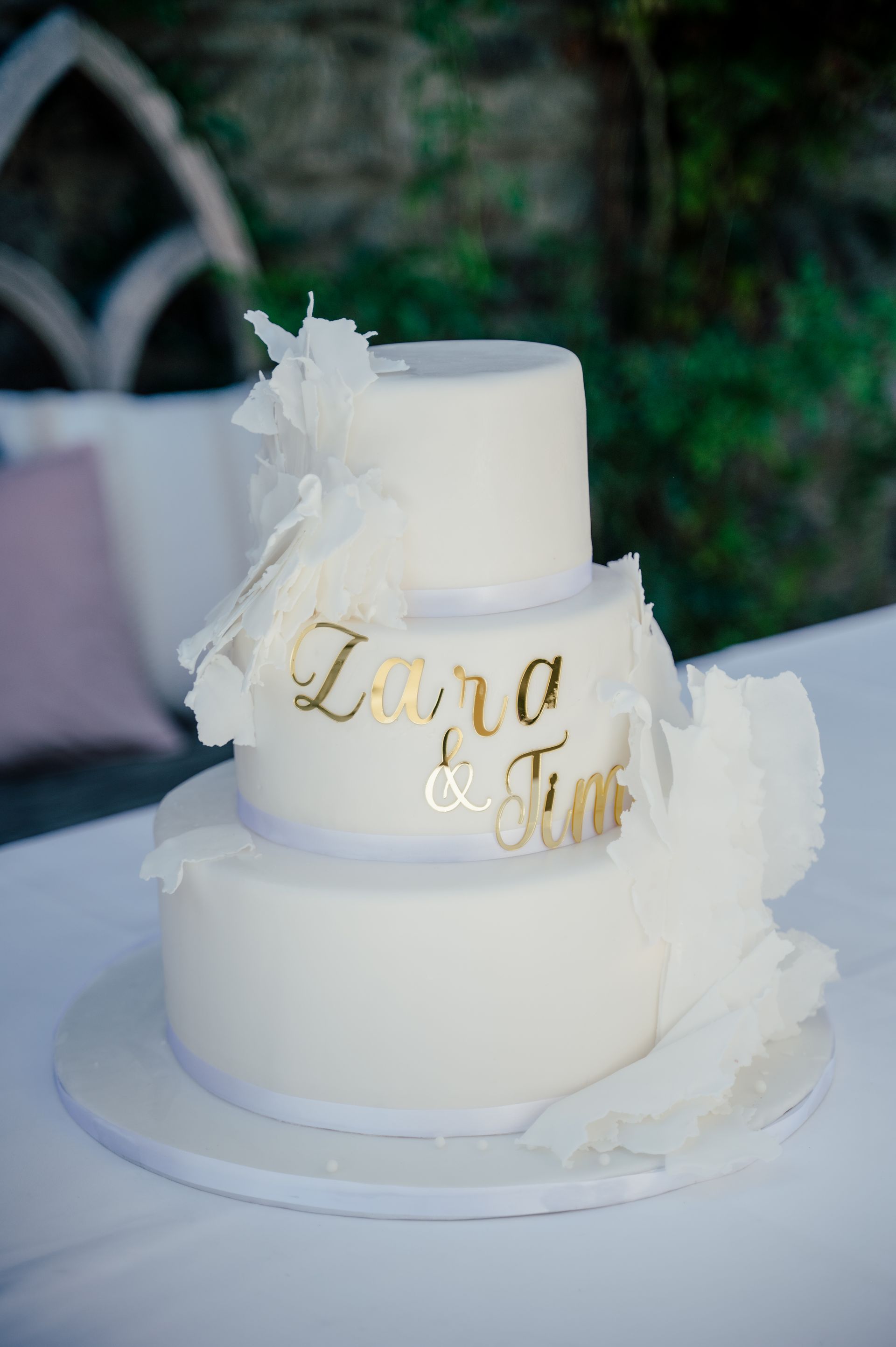A three-tier white wedding cake with ruffles and gold lettering reading “Zara & Tim” on an outdoor table.