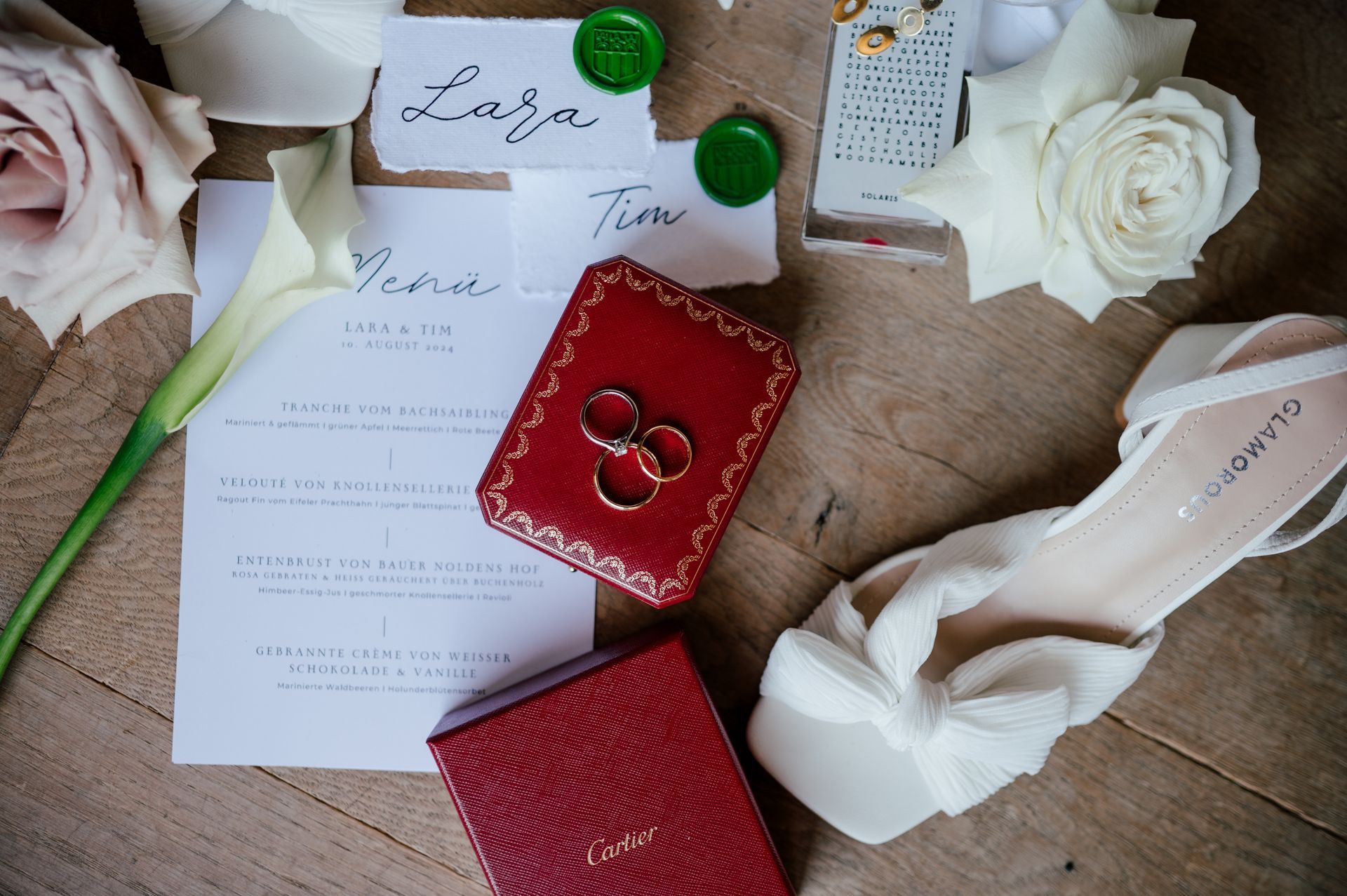 Wedding flat lay: Two red ring boxes containing wedding rings, invitation cards, a white shoe, and flowers on a wooden surface.