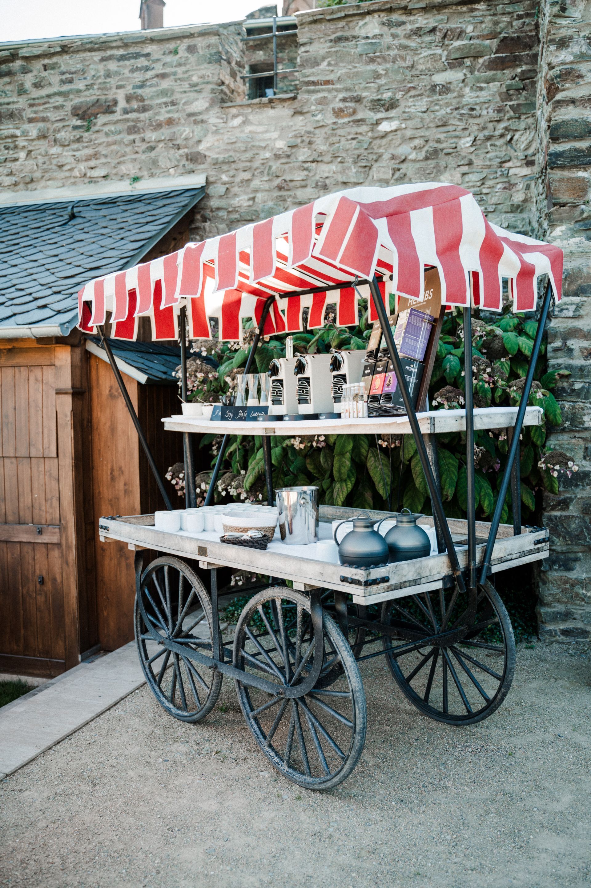 A wooden, four-wheeled serving cart with a red-and-white striped canopy, parked on a gravel path near a stone wall.