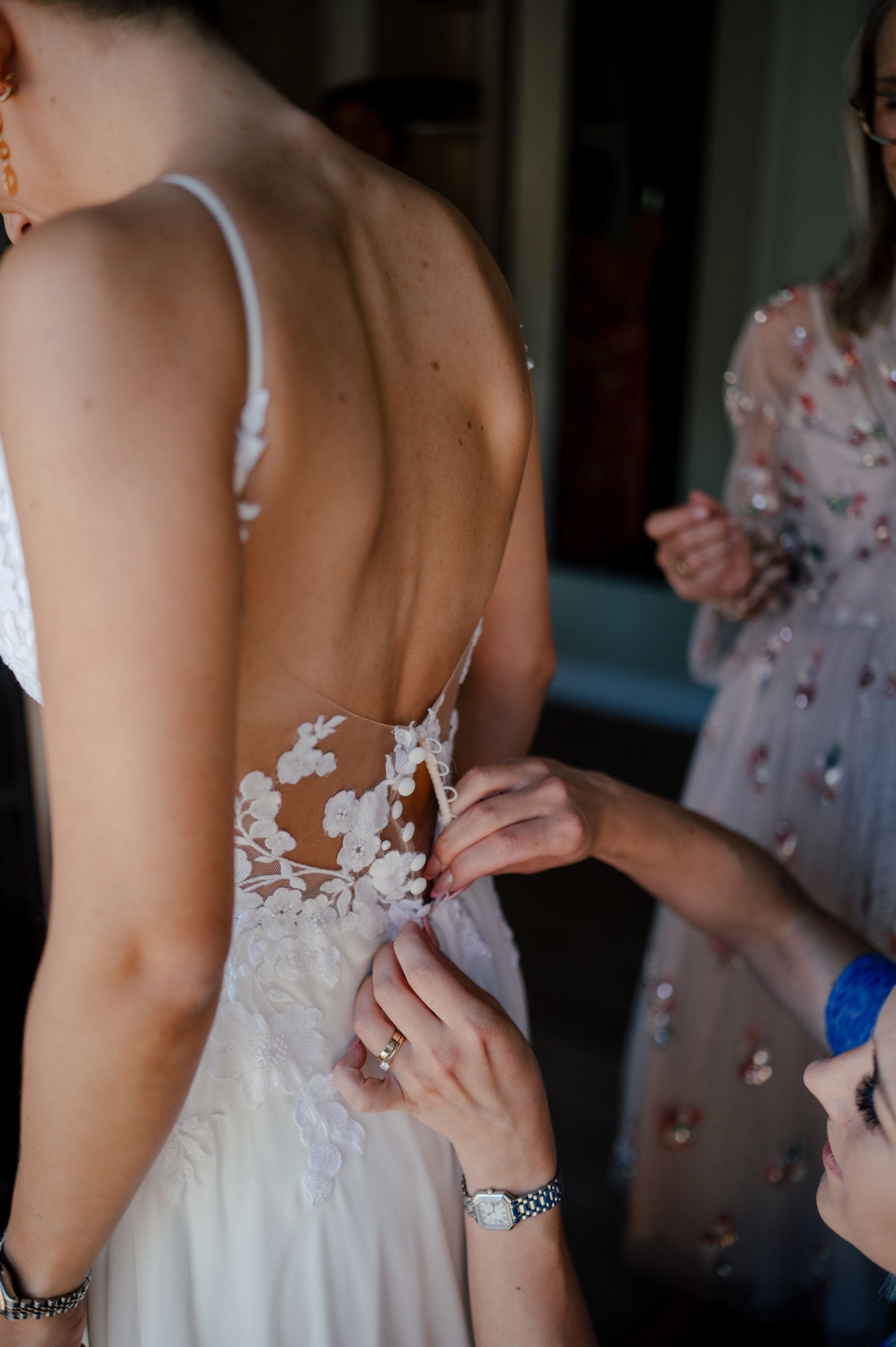 One person is adjusting the back of a lace wedding dress for another person, while a bystander watches in the background.