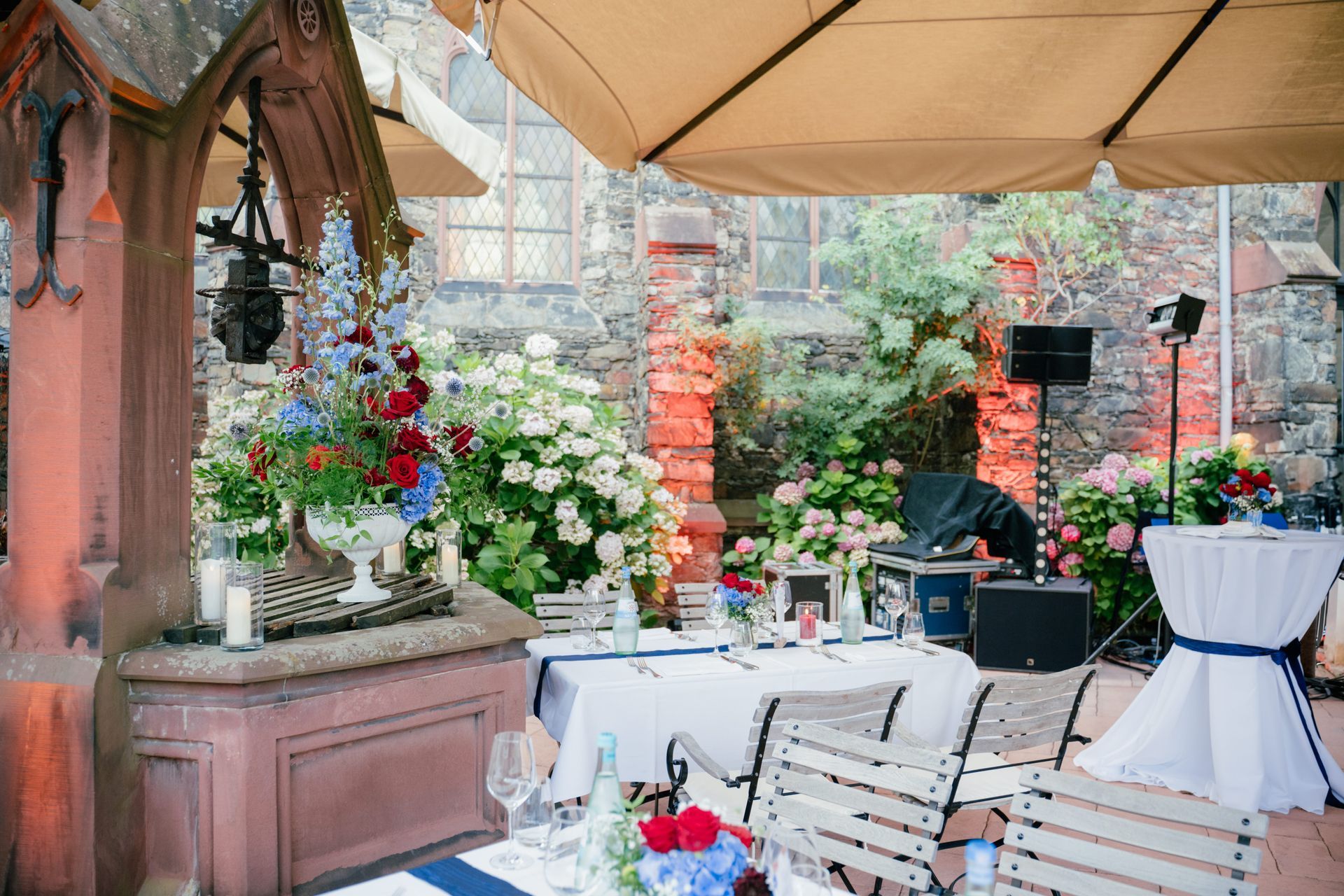 An outdoor garden event featuring stone walls, a decorative fountain as the centerpiece, tables with white tablecloths, and vibrant floral arrangements.